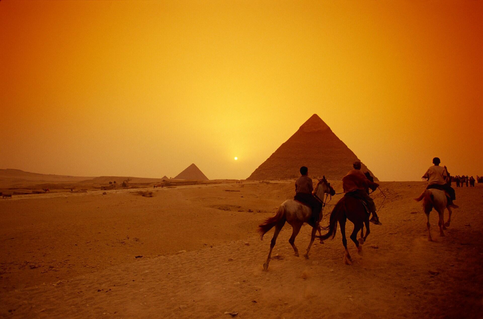 Riders on horses near pyramids with a sunset sky in Egypt.