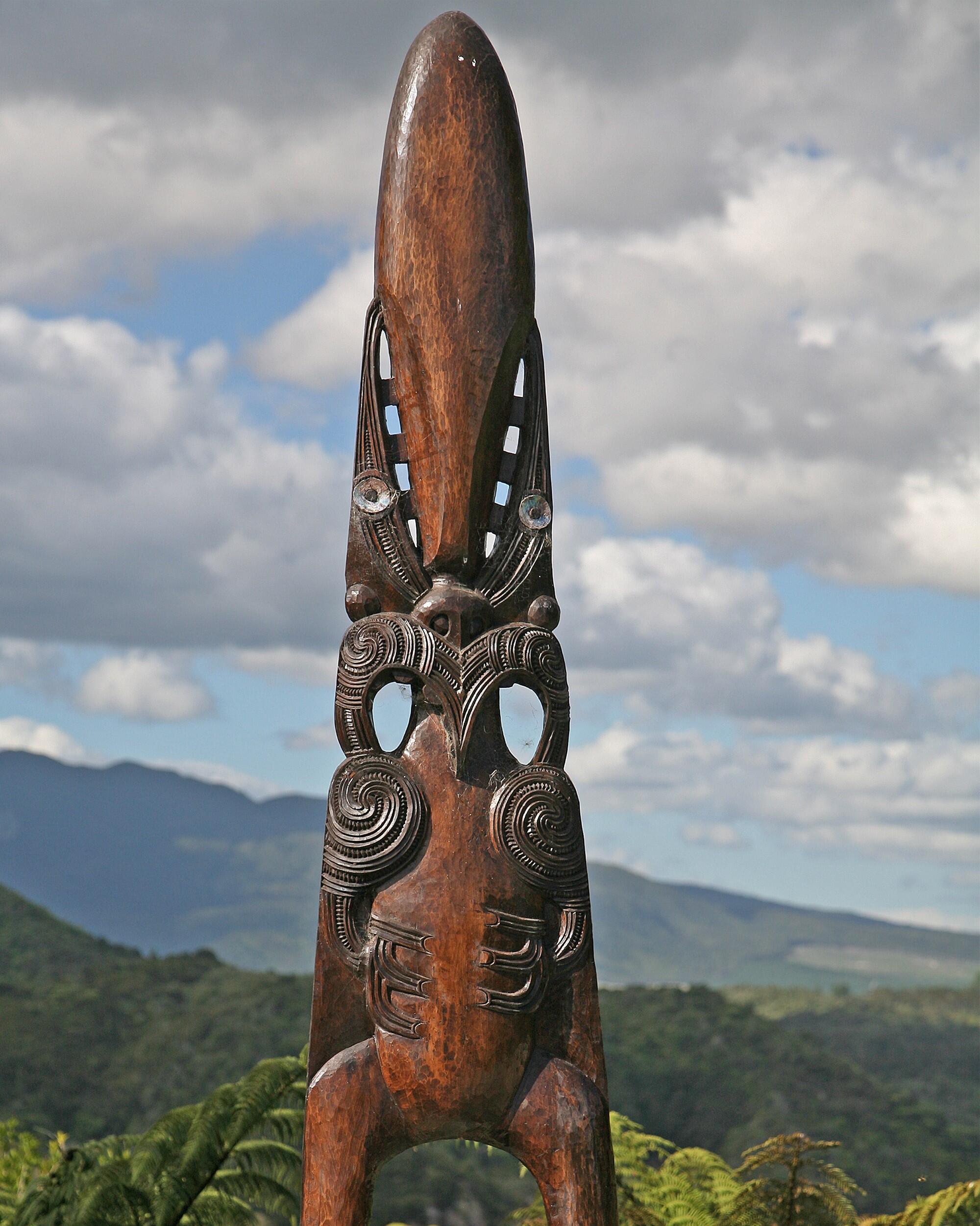 Tall carved wooden manaia figure standing outdoors against hills and sky.