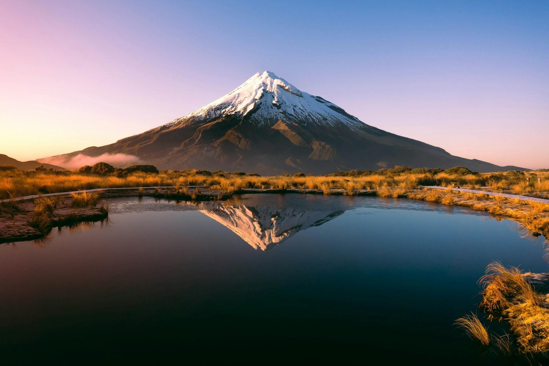 Mount Taranaki reflected in still water at sunrise, surrounded by wetlands and grass.