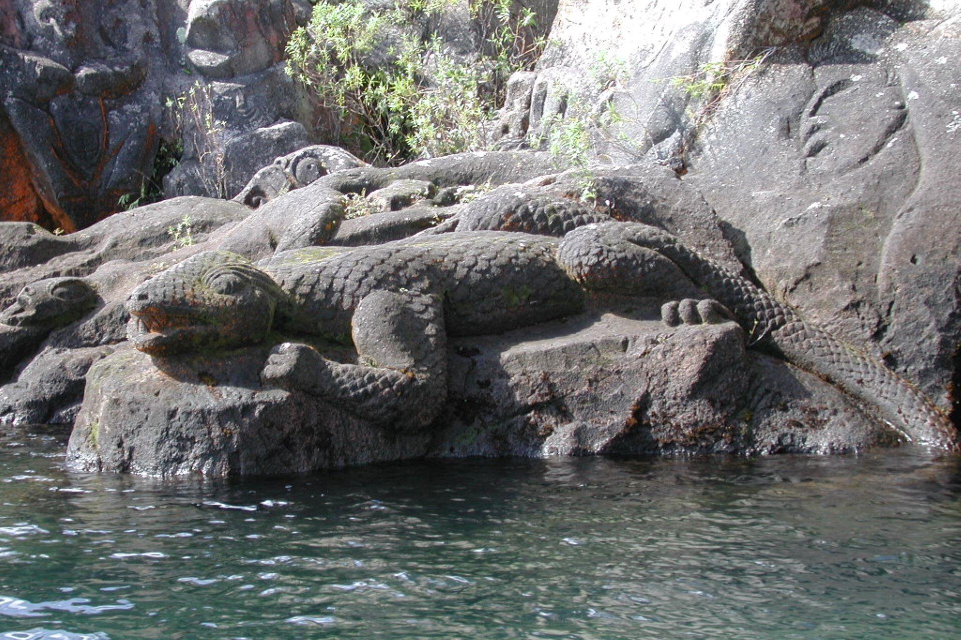 Stone carving of a taniwha-like creature resting among coastal rocks beside water.