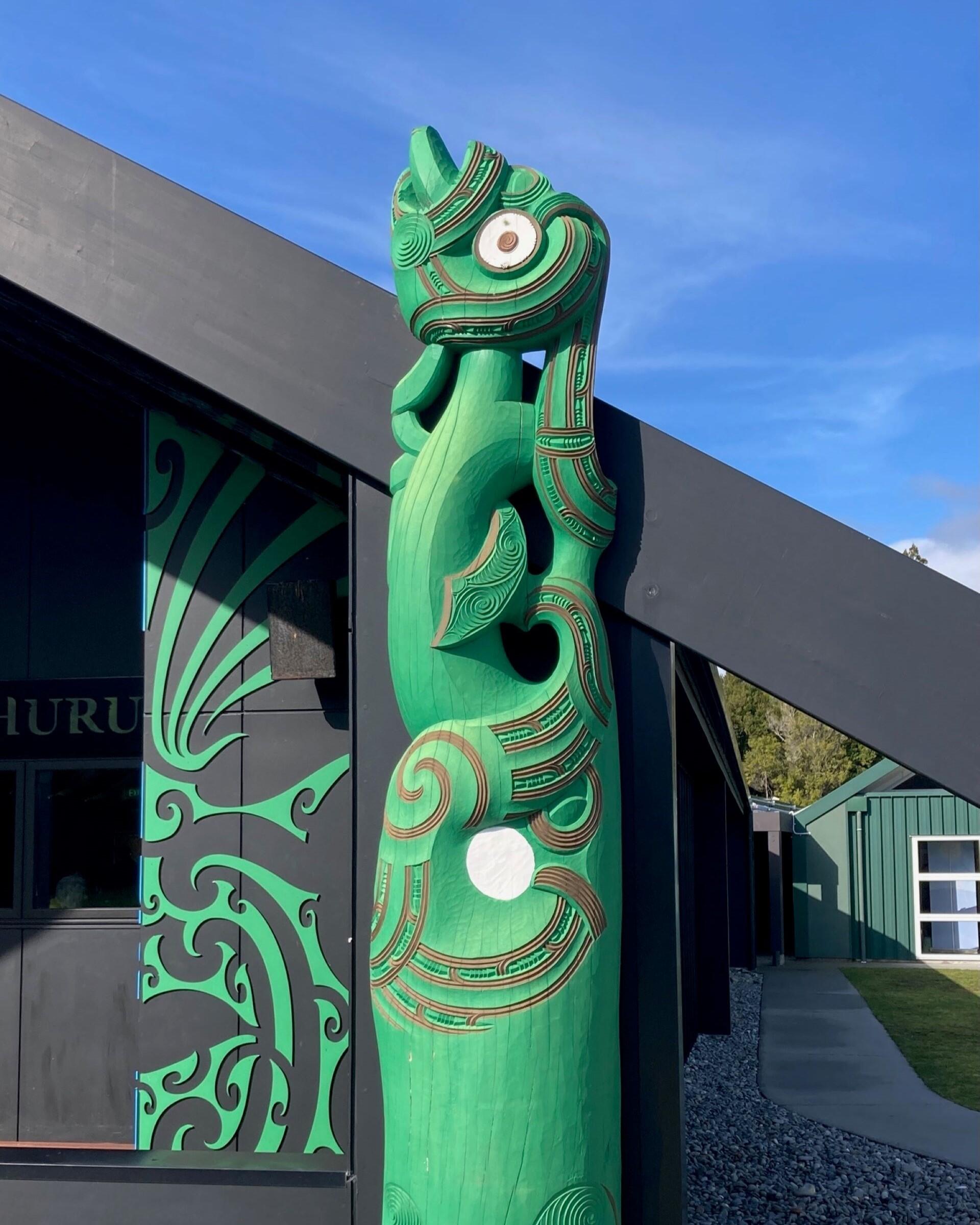 Green Māori wood carving depicting a taniwha figure outside a marae building.