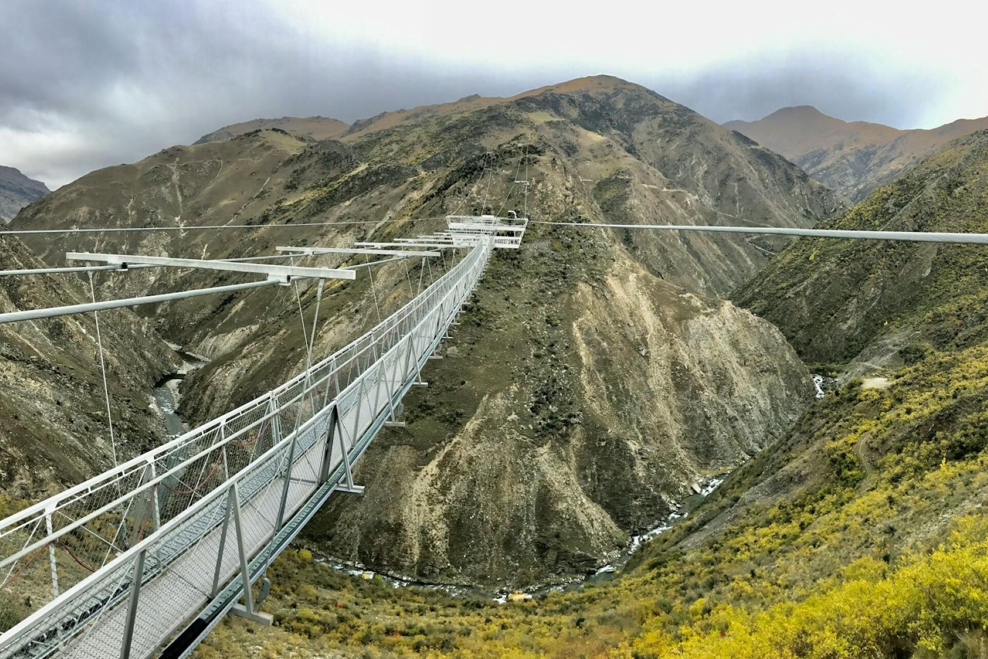 Suspension bridge spanning a rocky canyon with a river below and mountains in the background.