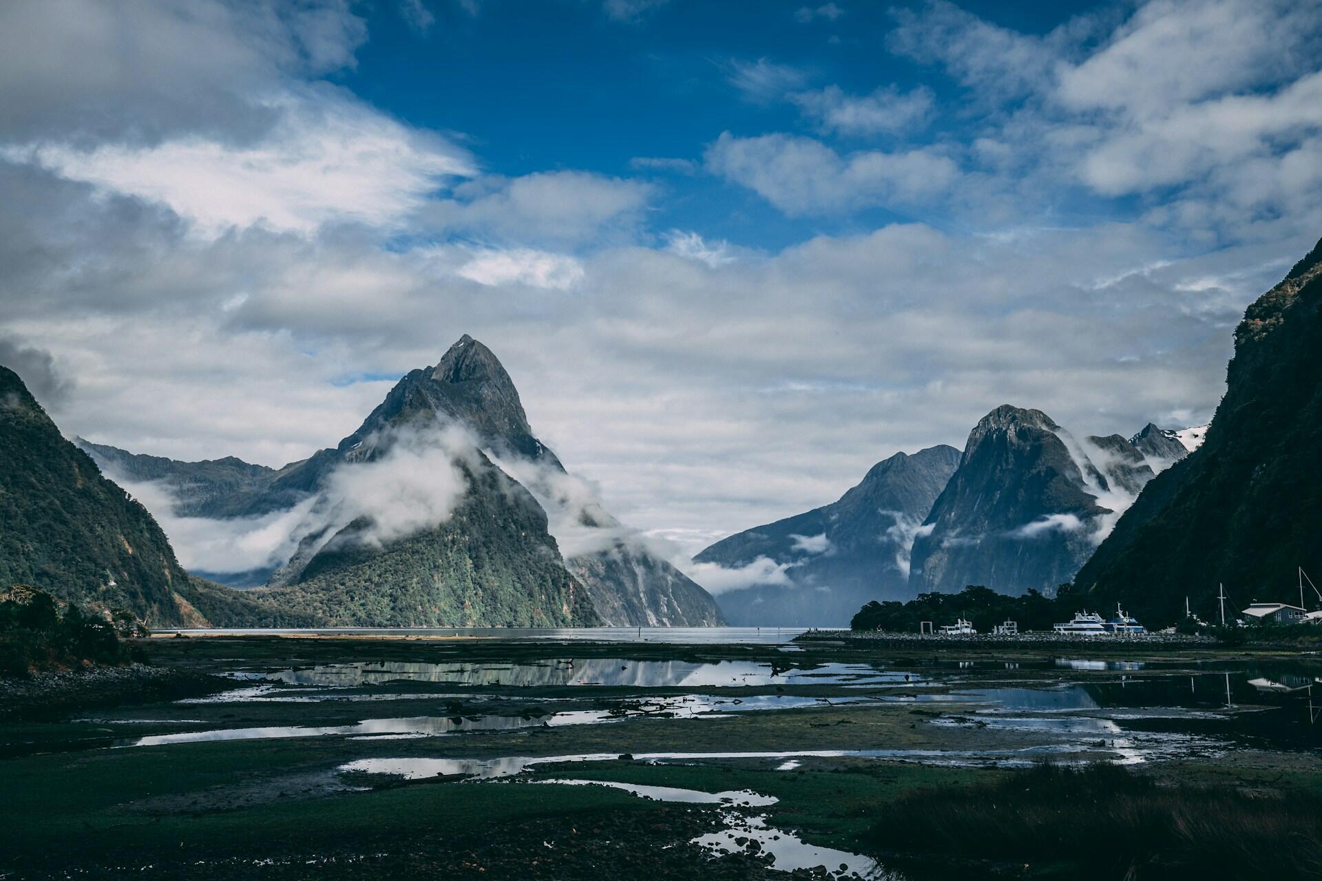 Milford Sound fjord with steep mountain peaks rising from the water, low clouds drifting around the cliffs, and reflections in the shallow foreground.