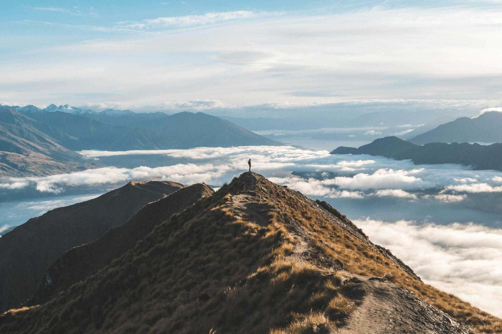 person standing on mountain ridge above clouds with distant mountains