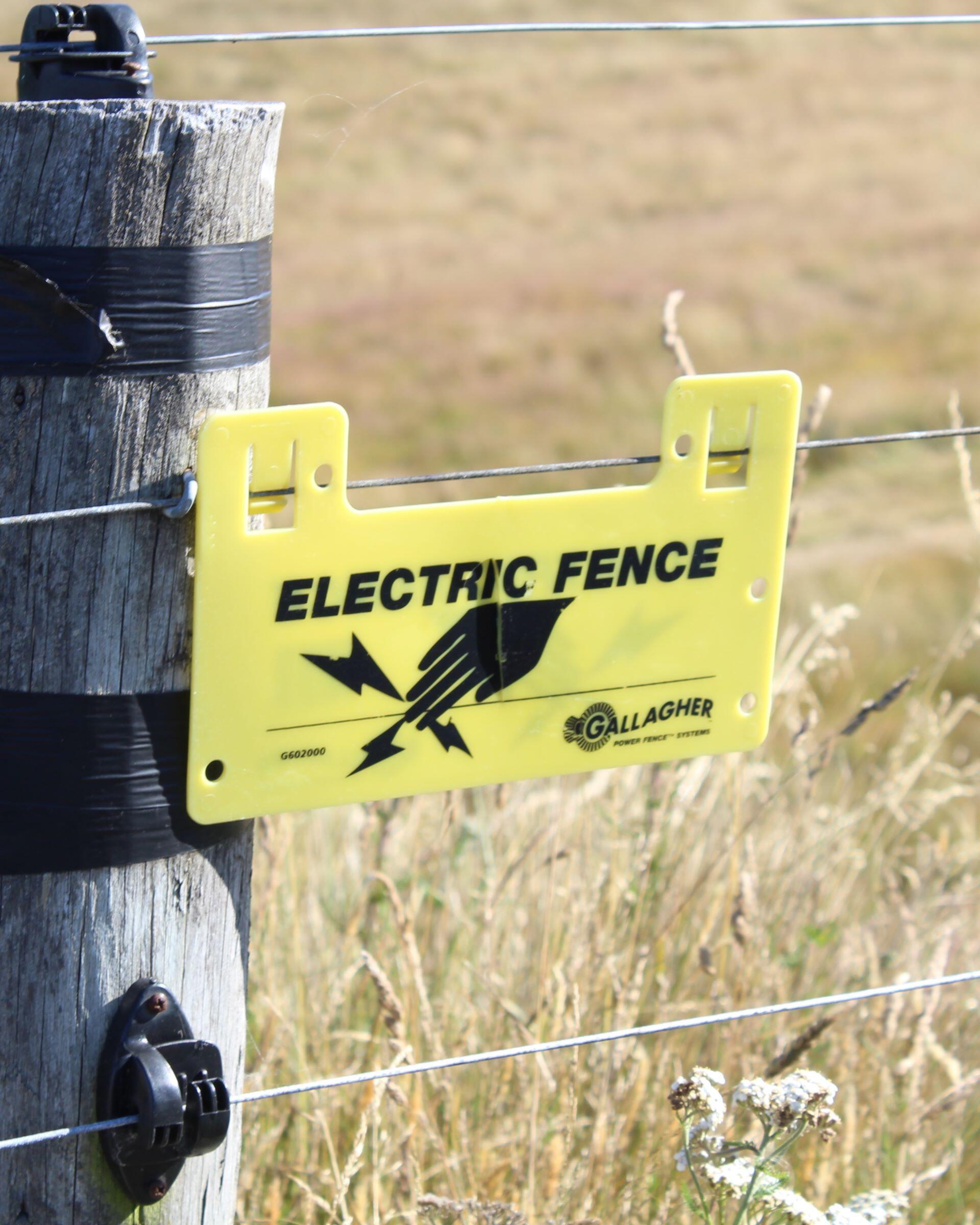 Yellow electric fence warning sign attached to a wire fence post in a field