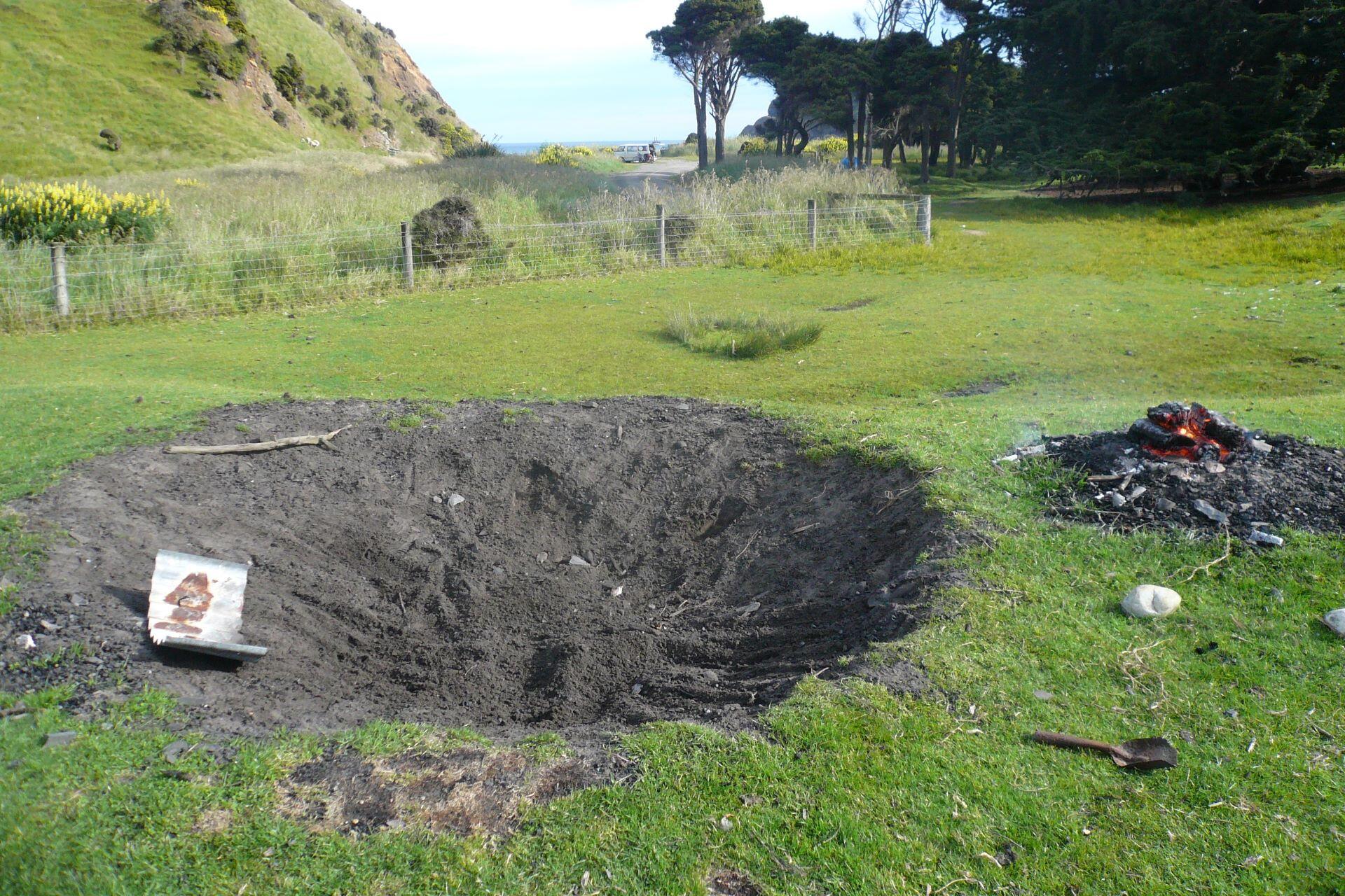 Large pit dug in grassy area with soil removed and small fire burning nearby