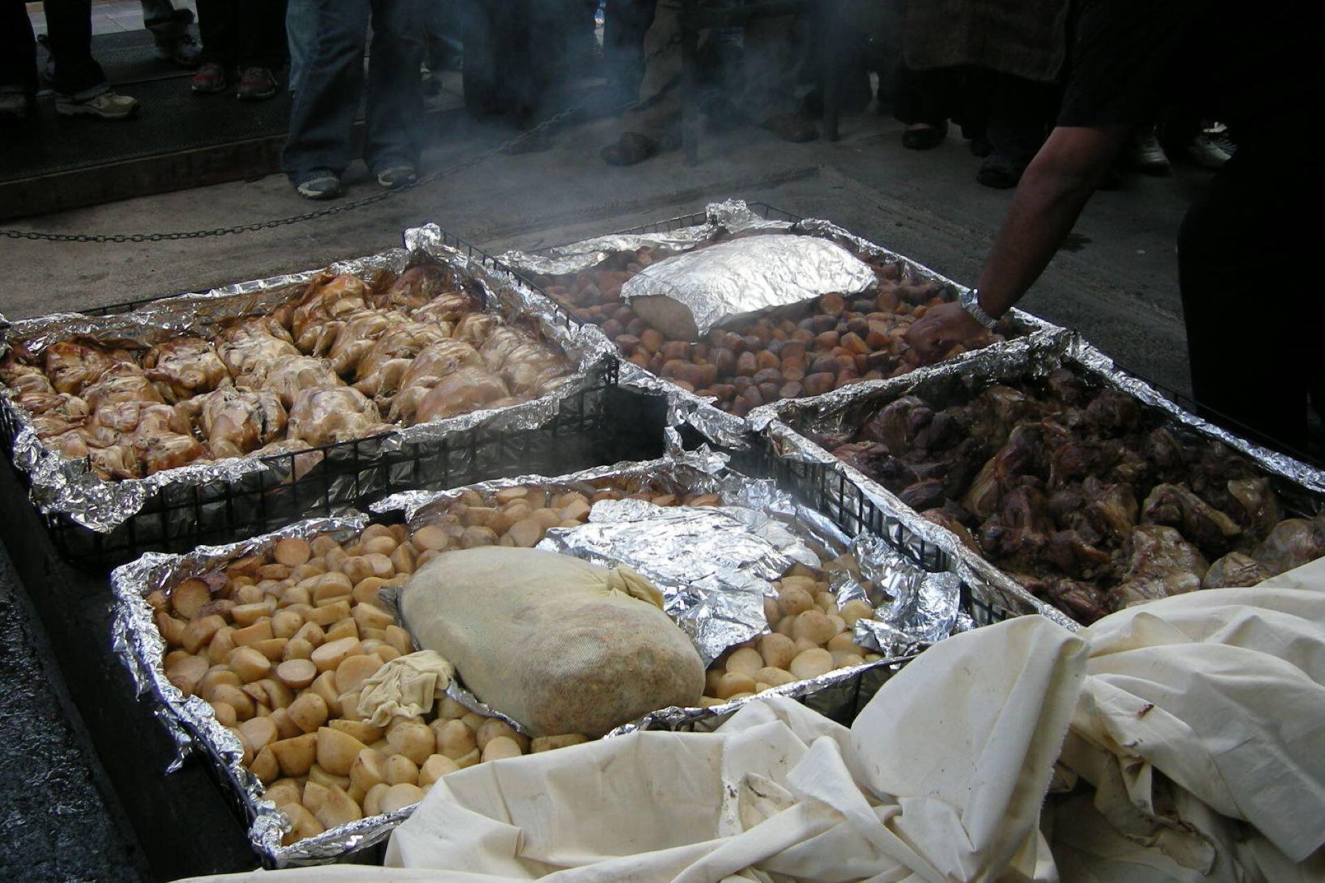 Multiple trays of meat and vegetables cooking in a traditional hāngī setup with steam rising