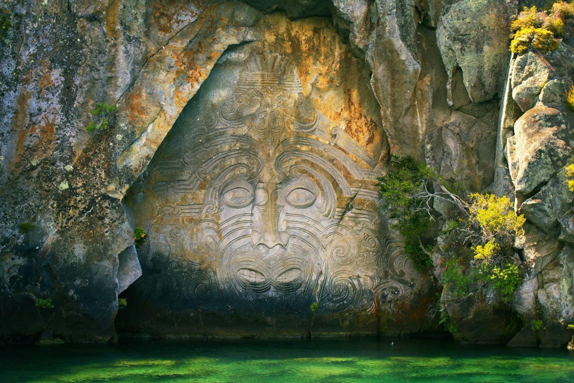 large carved face in rock cliff above water surrounded by vegetation