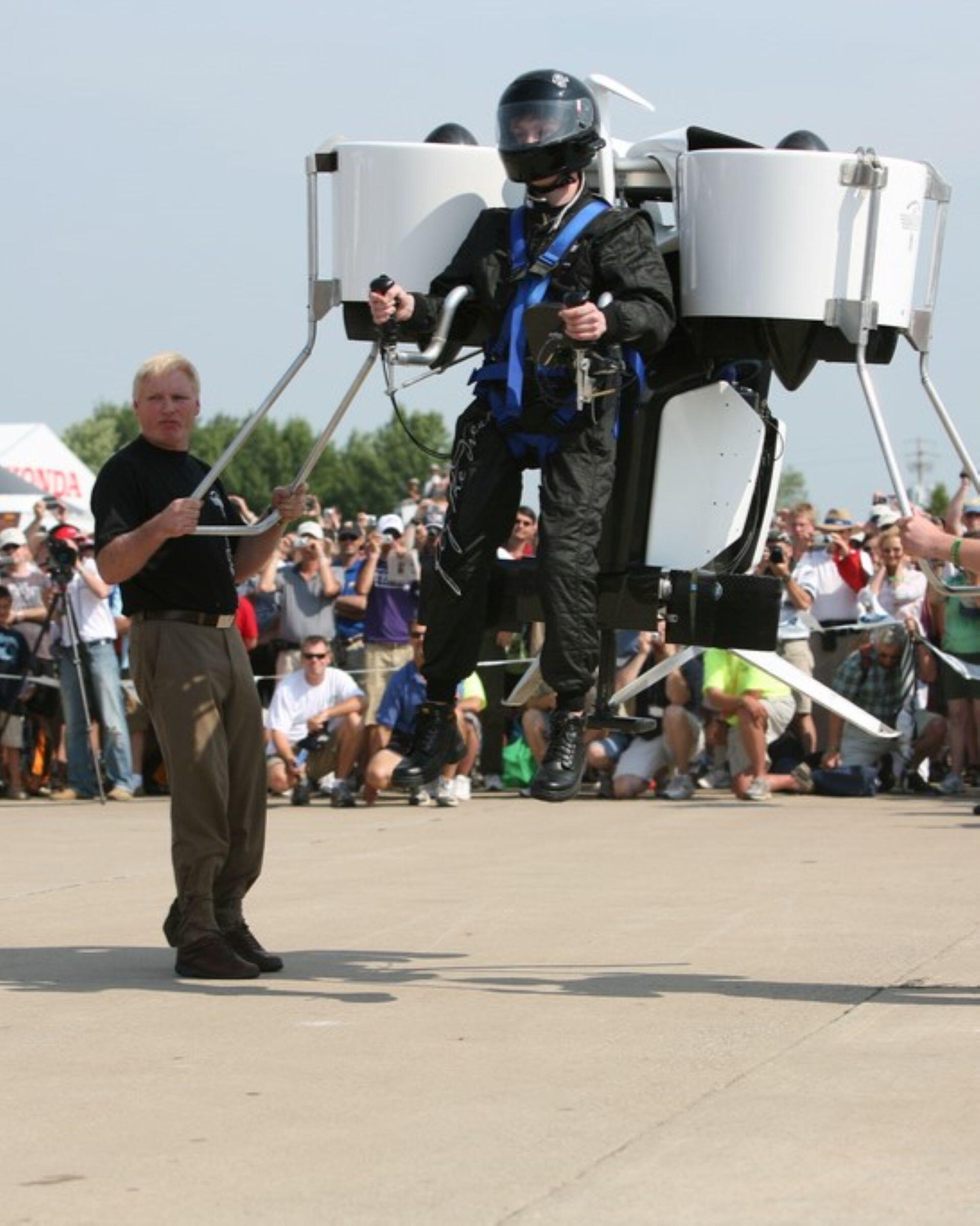 Person wearing a jetpack device hovering above the ground with a crowd in the background