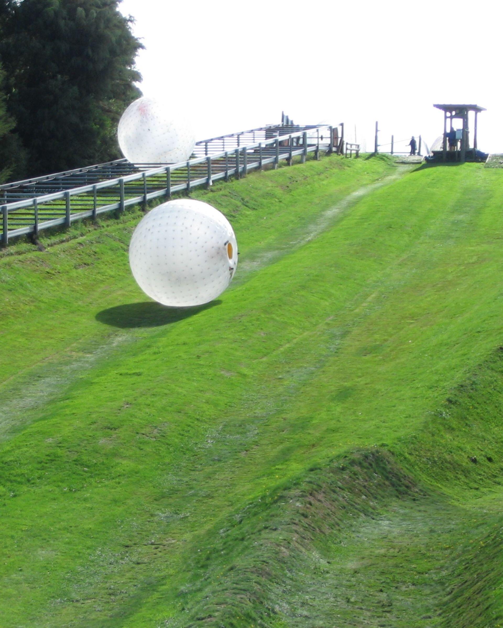 Two large inflatable zorb balls rolling down a grassy hill with a track