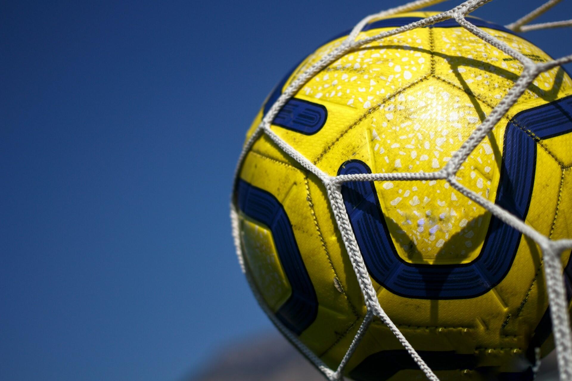 Yellow football caught in a goal net against a blue sky.