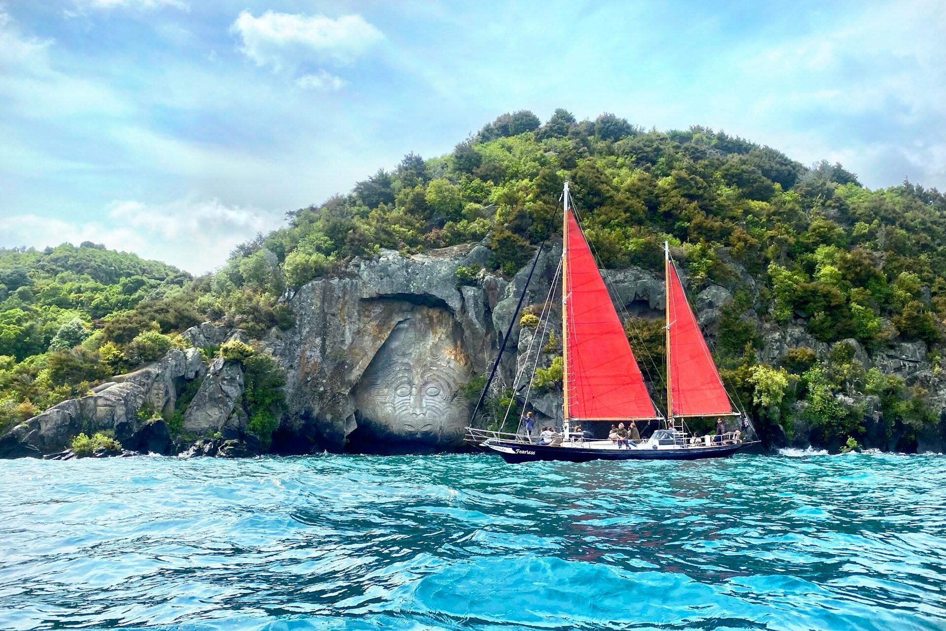 A red-sailed boat on blue water beside a rocky green shoreline with a large rock carving