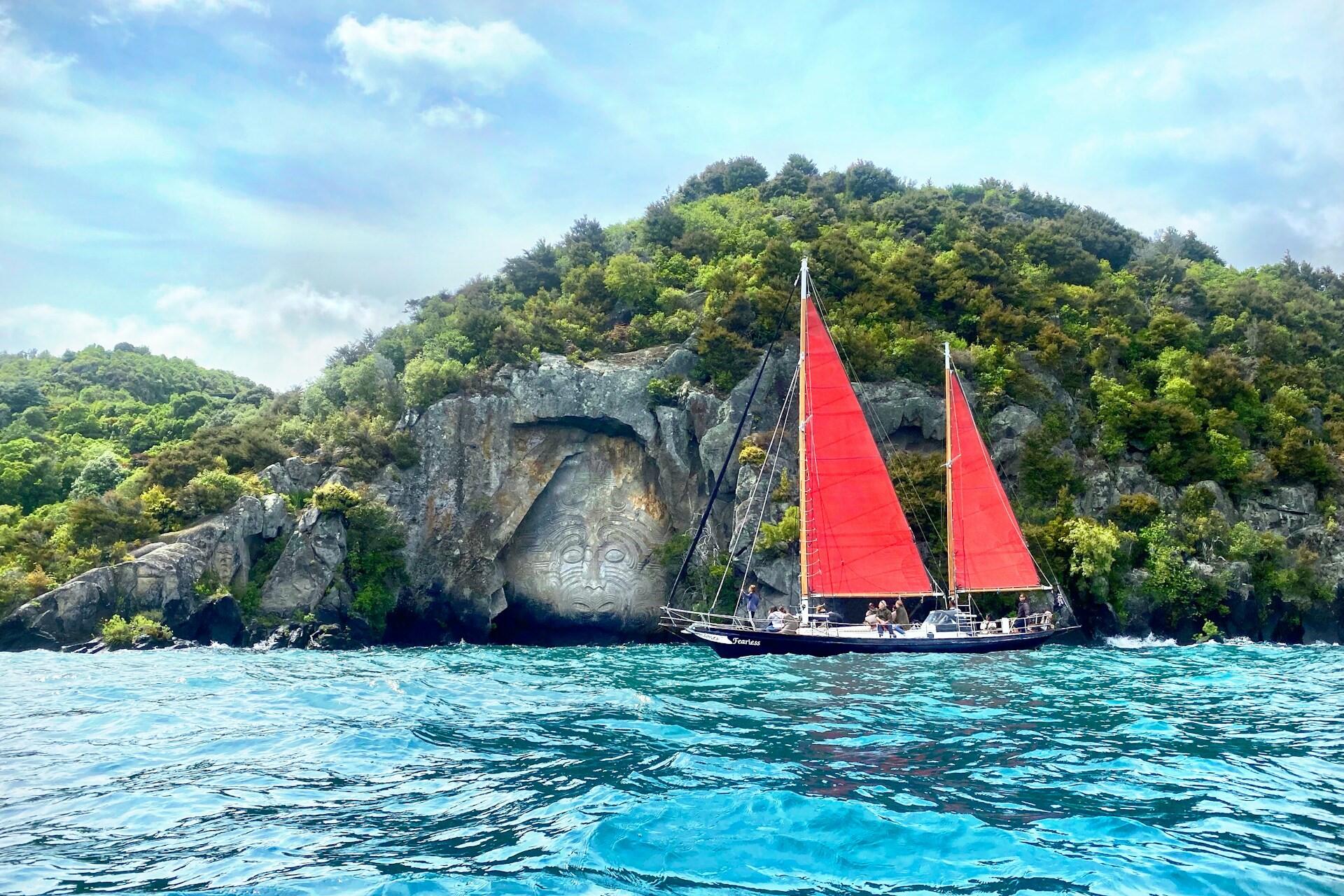 Māori rock carving beside the water in New Zealand with a red-sailed boat