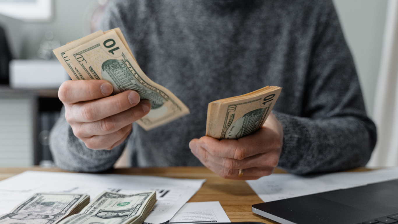 Persona contando billetes de 10 dólares sobre una mesa con documentos, dinero en efectivo y una laptop.