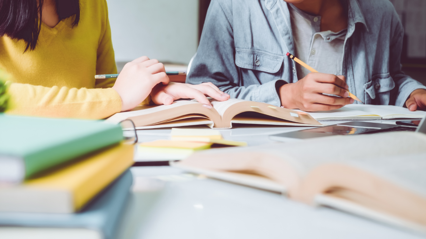 Dos estudiantes estudiando juntos en una mesa con libros abiertos, tomando notas con lápices y rodeados de material de estudio.