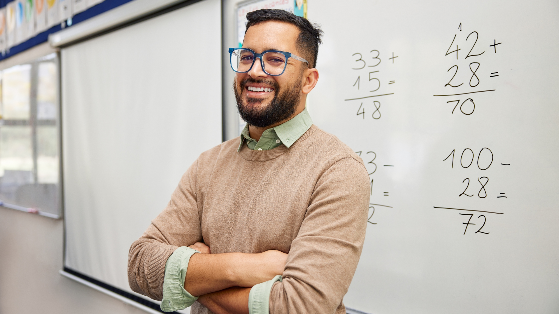 Profesor de matemáticas sonriente frente a un pizarrón con operaciones aritméticas en un aula.