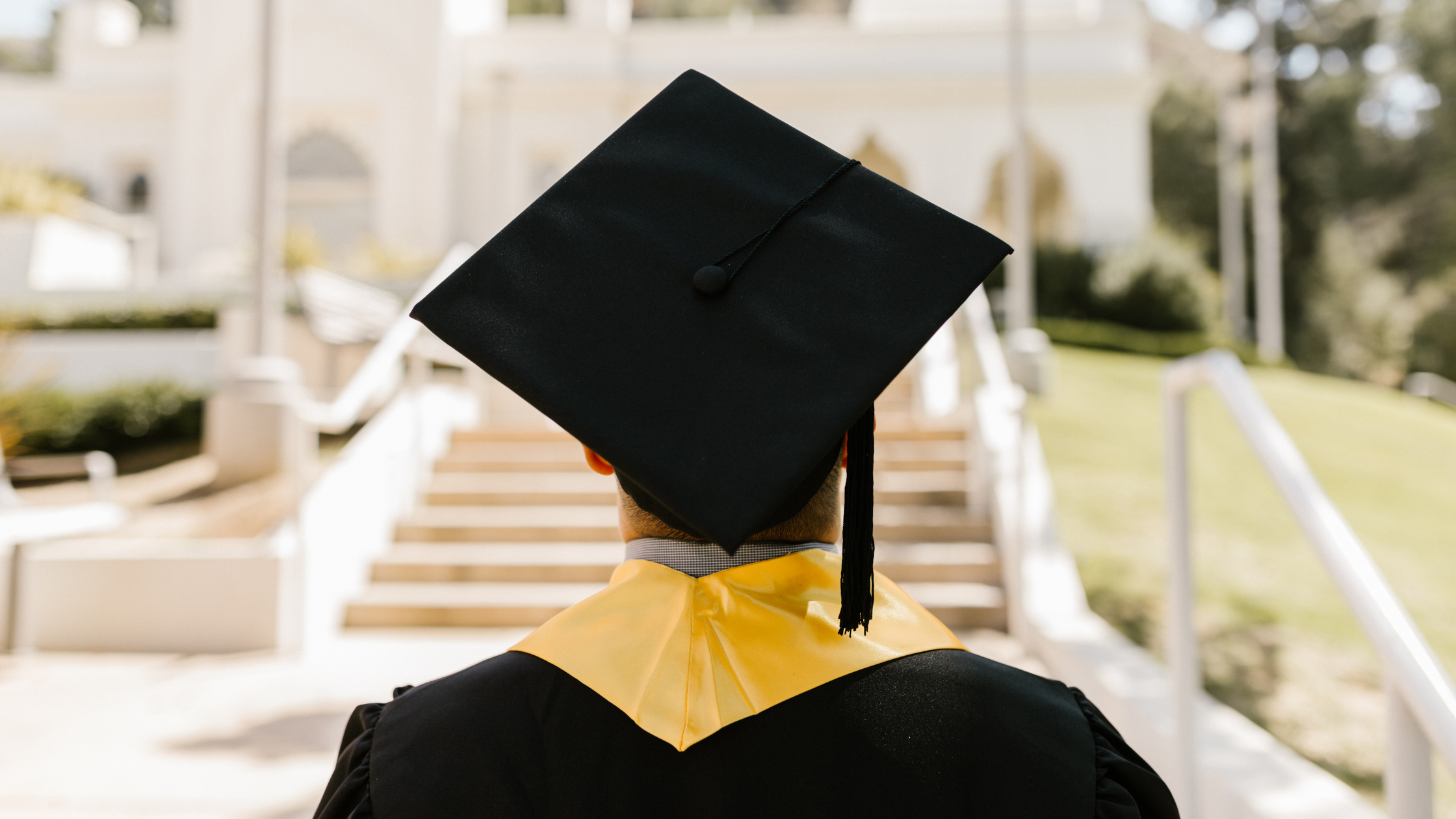 Graduado con toga y birrete negro de espaldas, mirando hacia una escalera en un campus universitario iluminado por el sol.