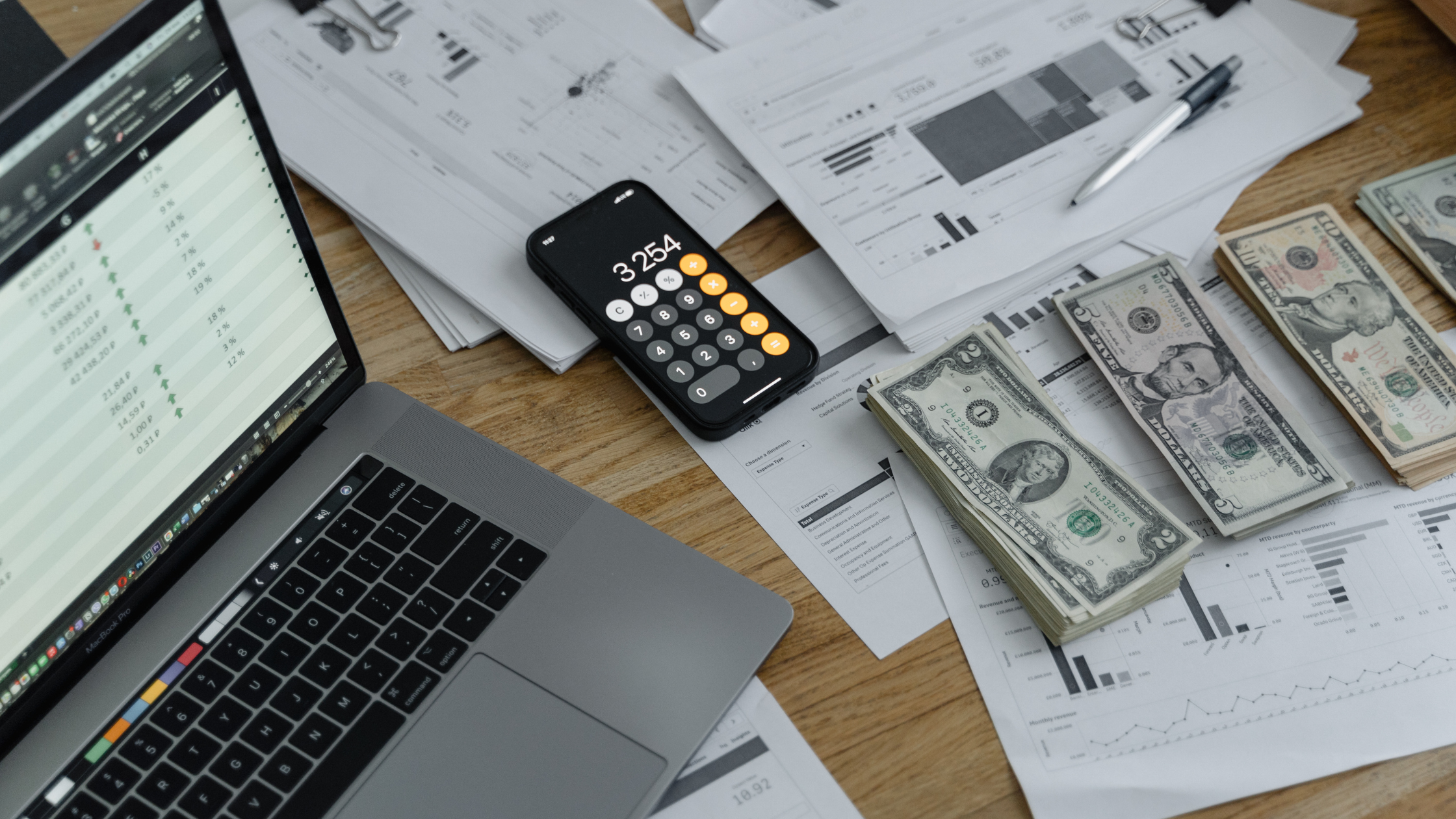 Escritorio con laptop, calculadora en un teléfono, pilas de billetes en dólares y documentos financieros con gráficos y estadísticas.