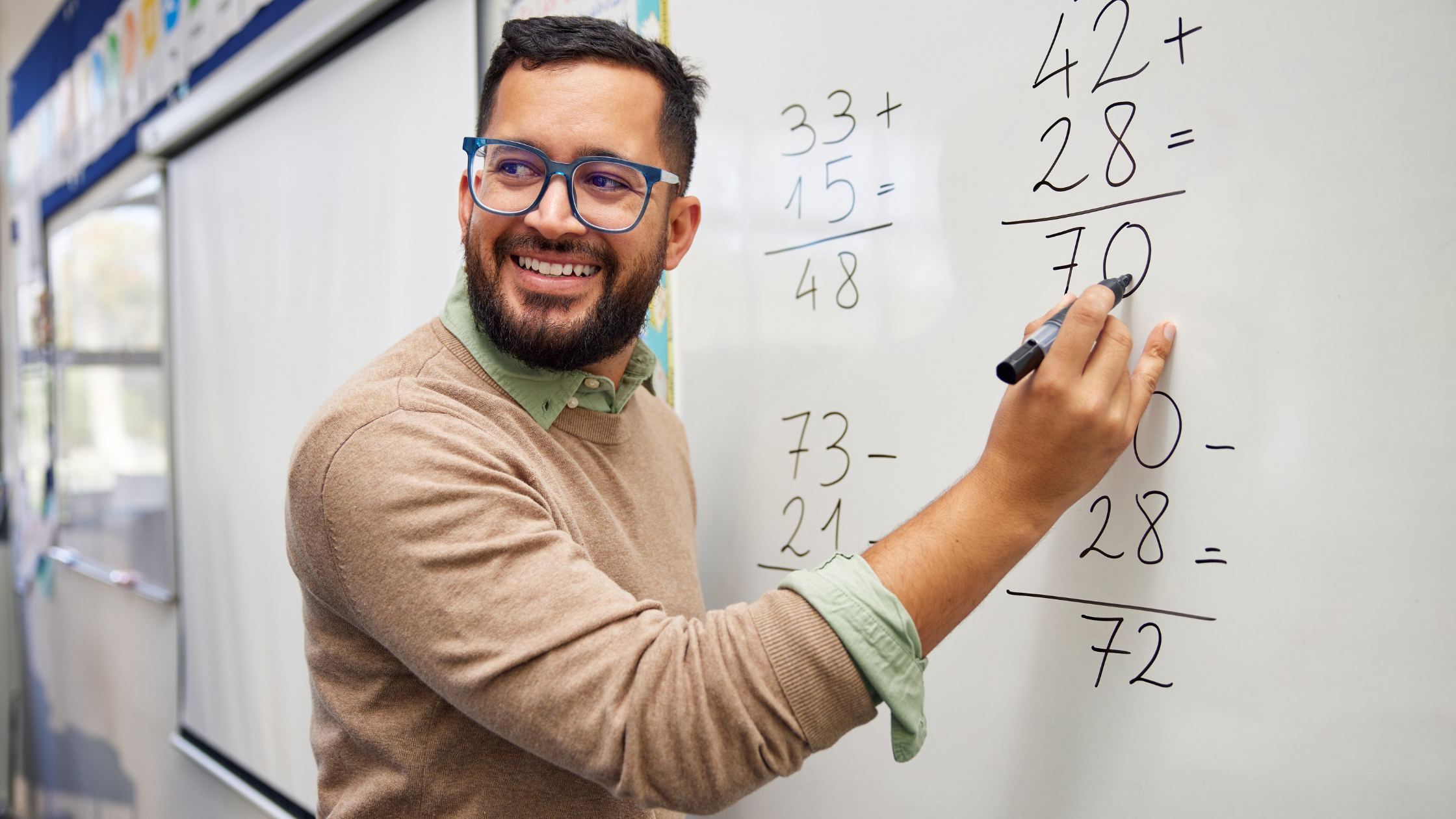 Profesor de matemáticas sonriente escribiendo operaciones en un pizarrón dentro de un aula.