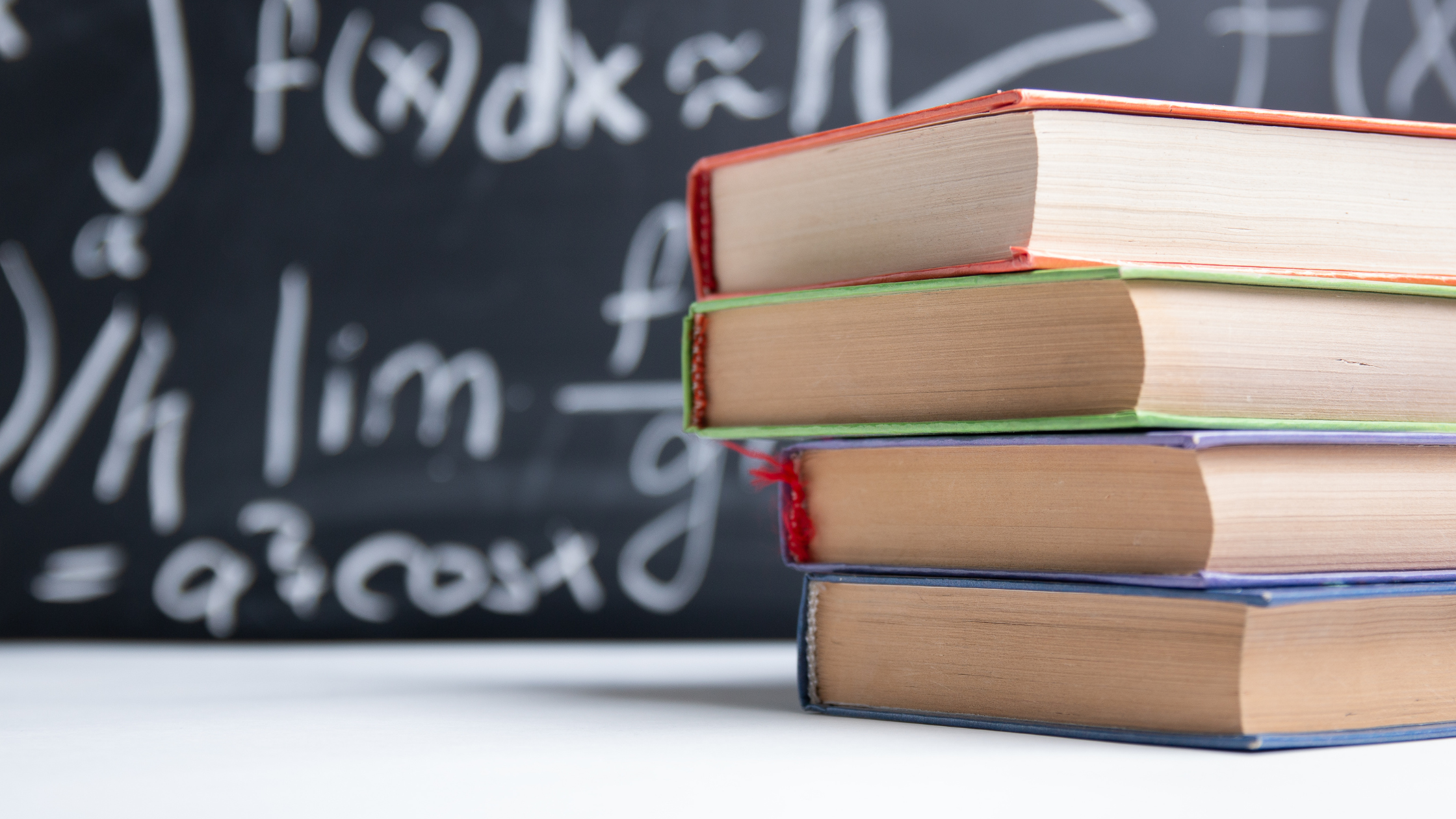 Pila de libros frente a un pizarrón con ecuaciones matemáticas escritas en tiza.