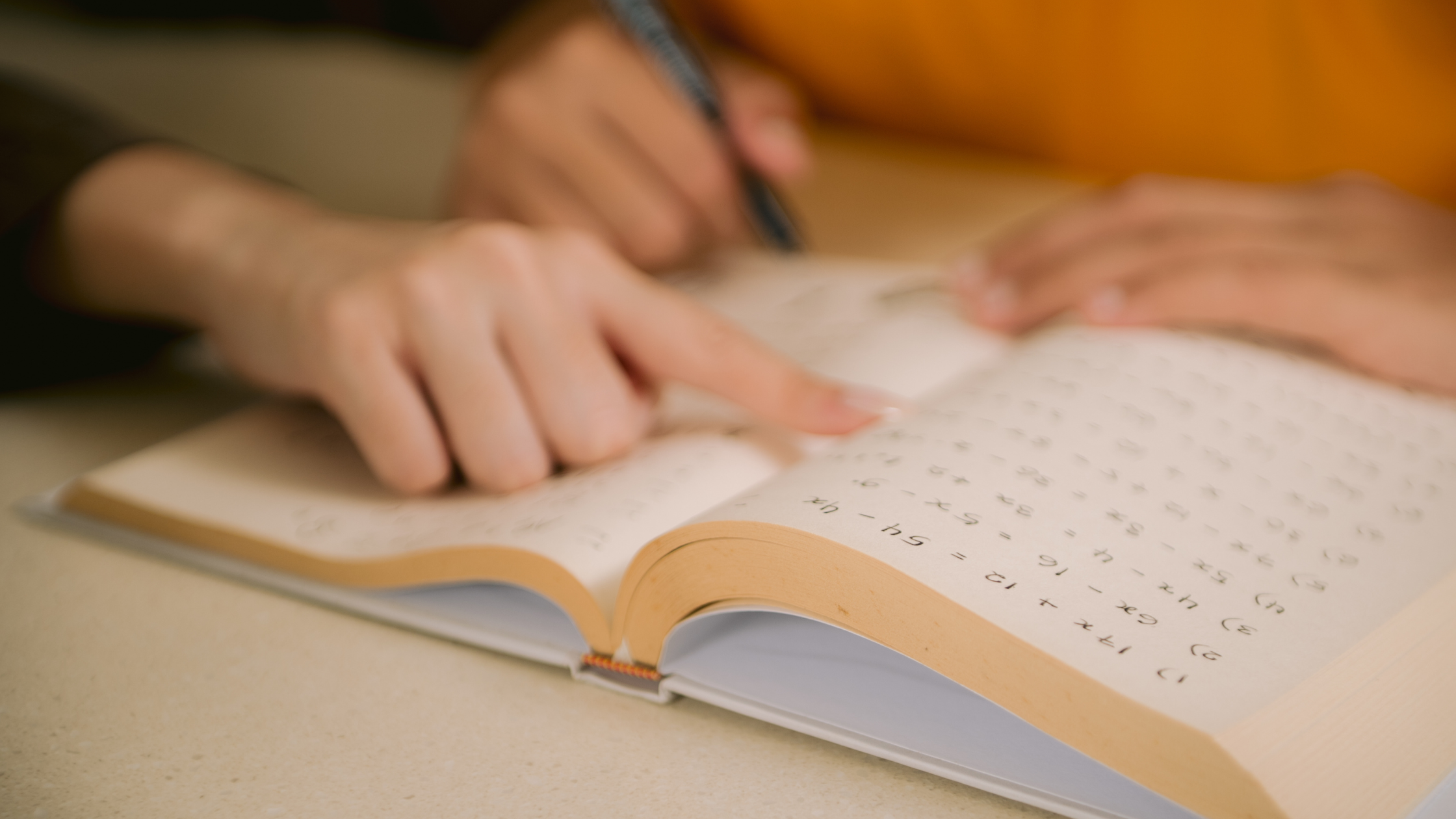 Primer plano de dos personas estudiando un libro de matemáticas, con una mano señalando una ecuación y otra escribiendo con un lápiz.