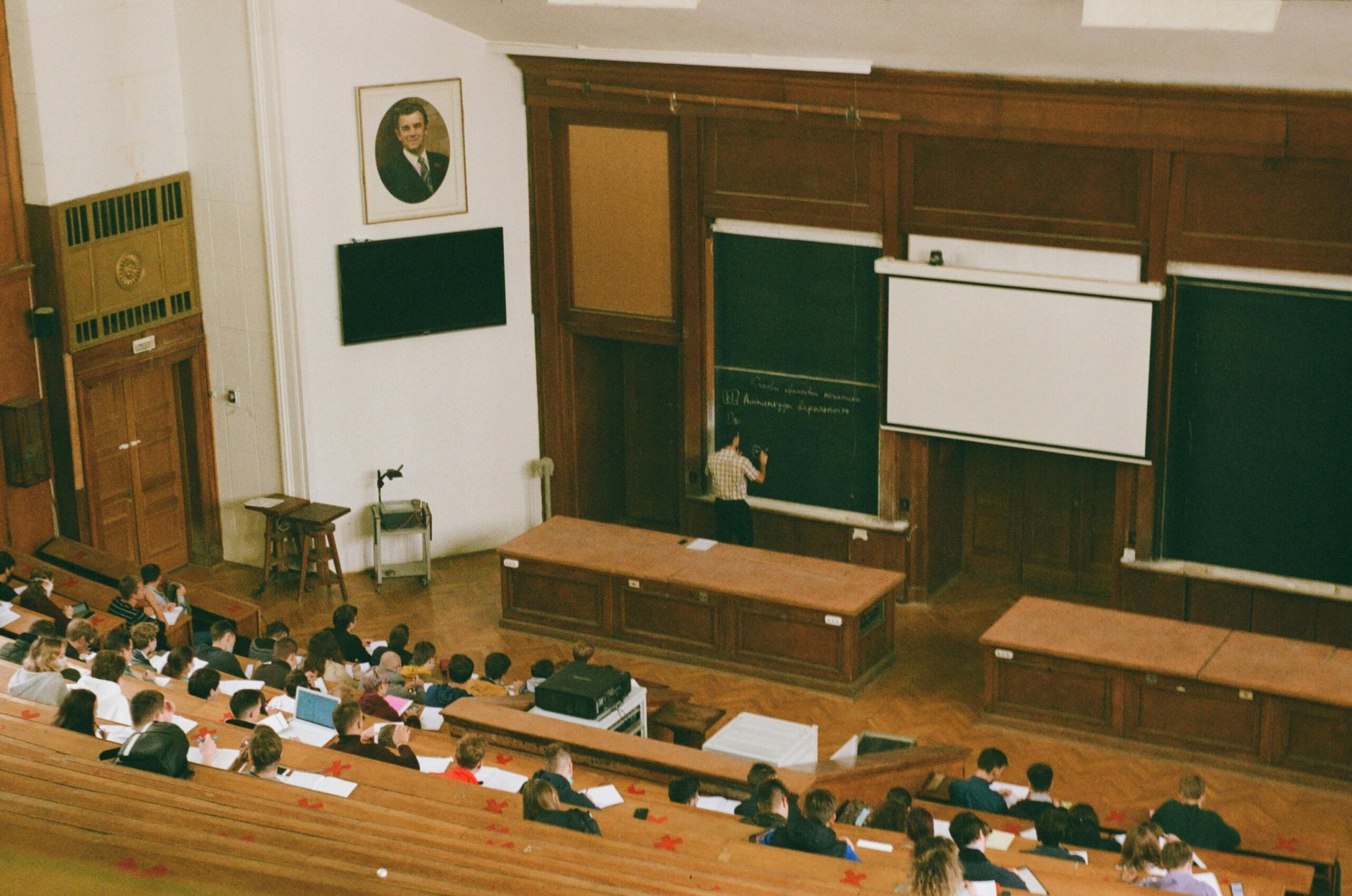 Vista panorámica de un auditorio universitario con estudiantes tomando notas mientras un profesor escribe en la pizarra.