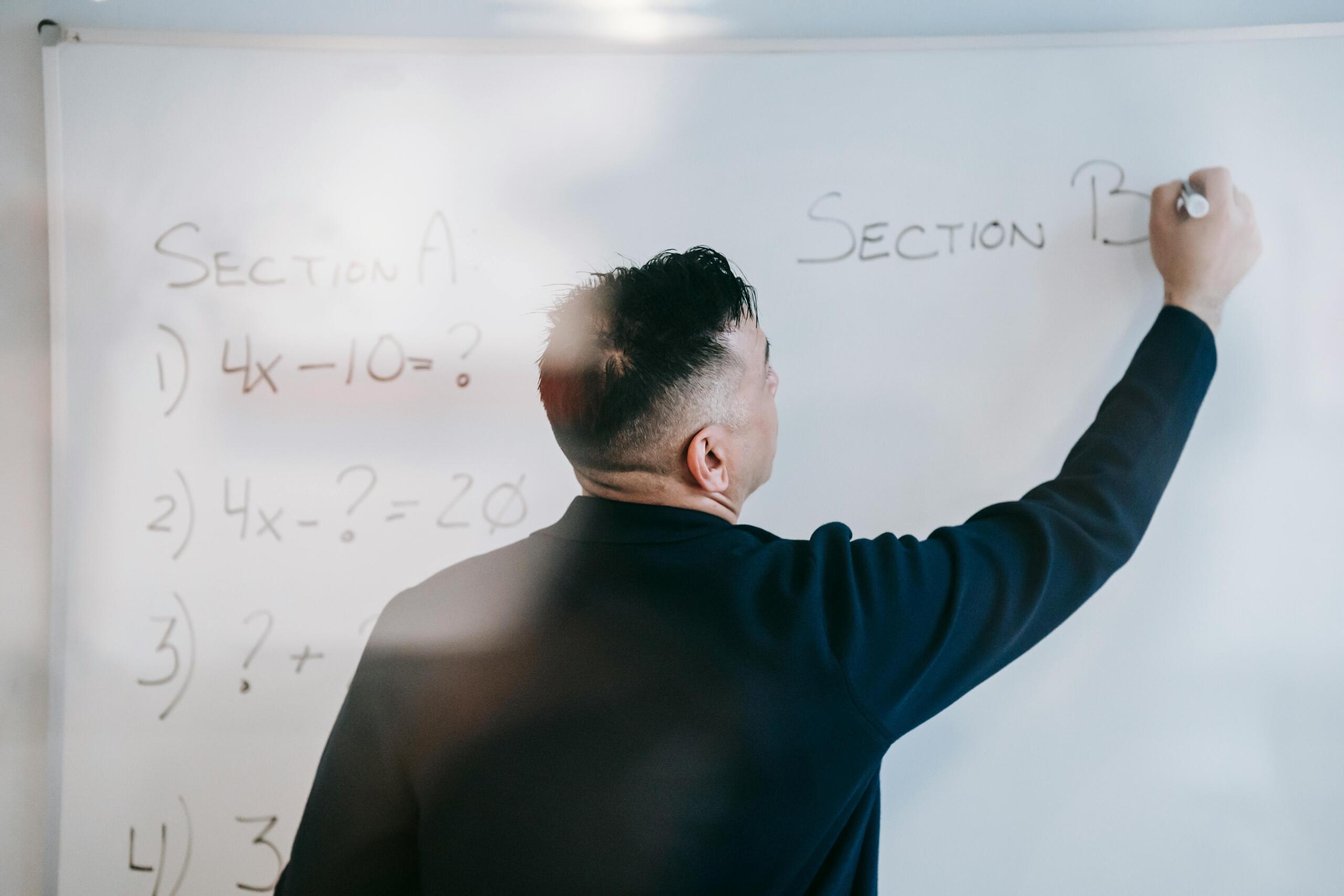 Profesor escribiendo secciones de un examen de álgebra en una pizarra blanca con operaciones de primer grado.