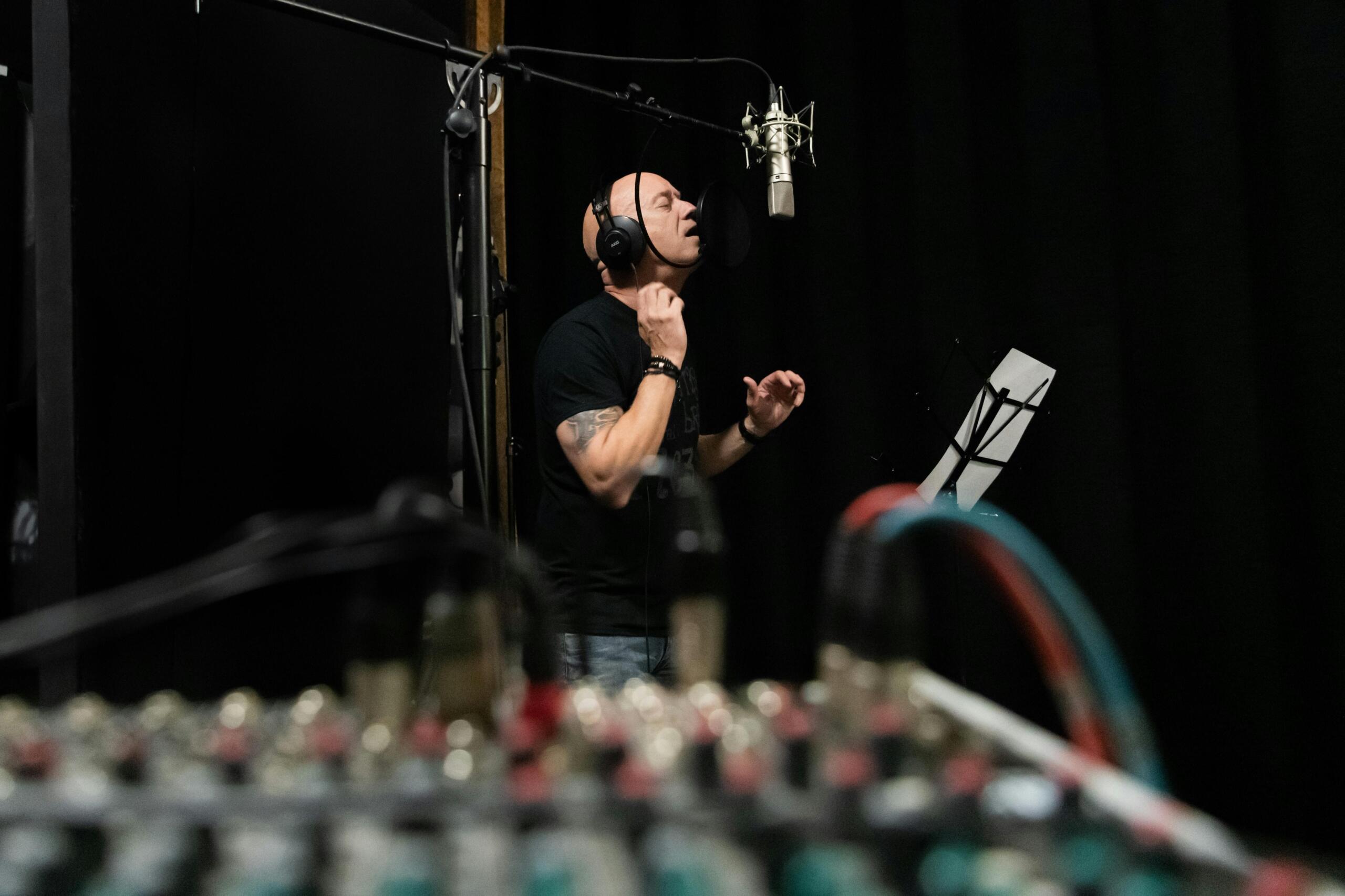 Vocalista masculino grabando en cabina con micrófono suspendido y consola de audio en primer plano.