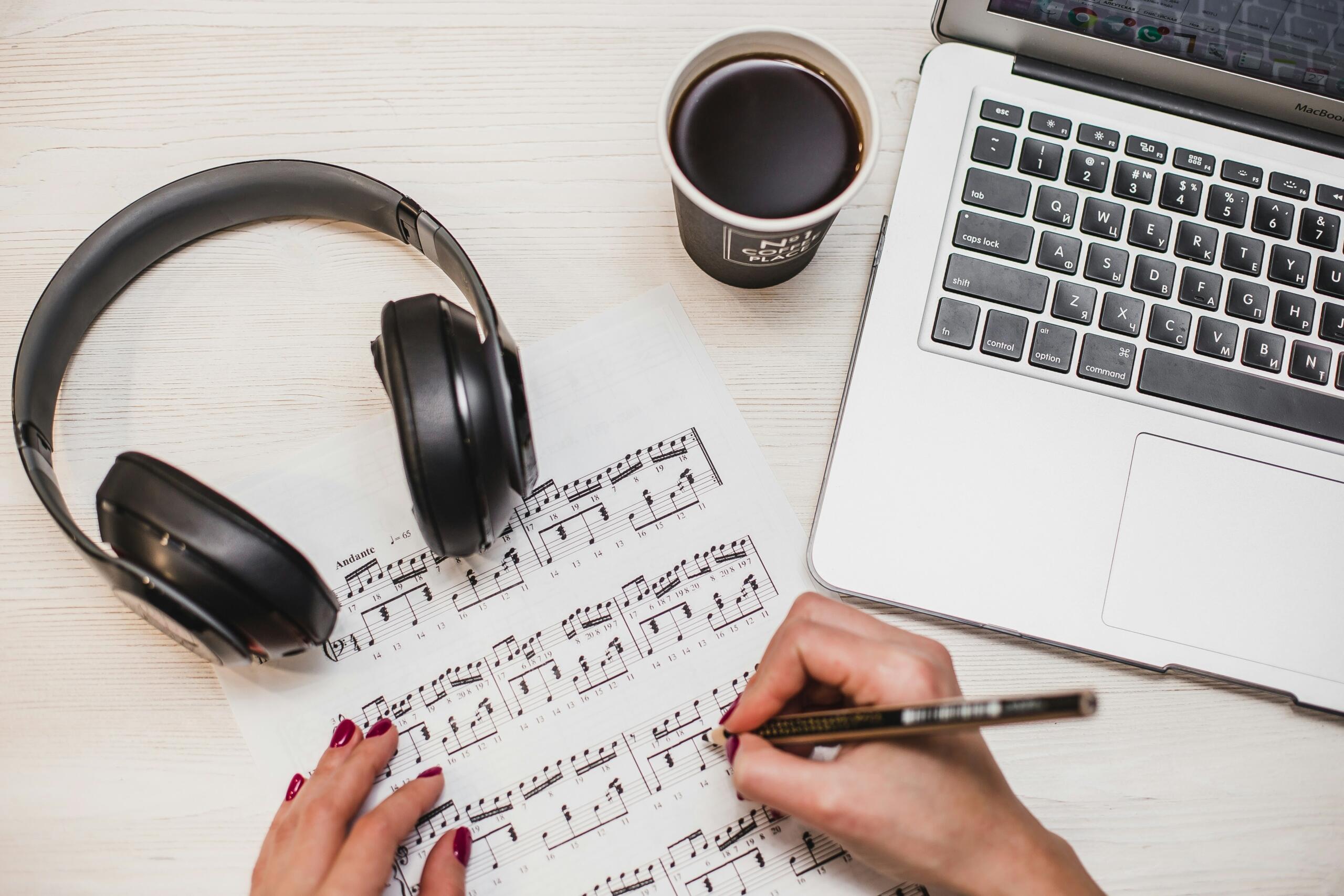 Mano escribiendo notas musicales junto a auriculares y laptop, composición y análisis vocal en proceso.