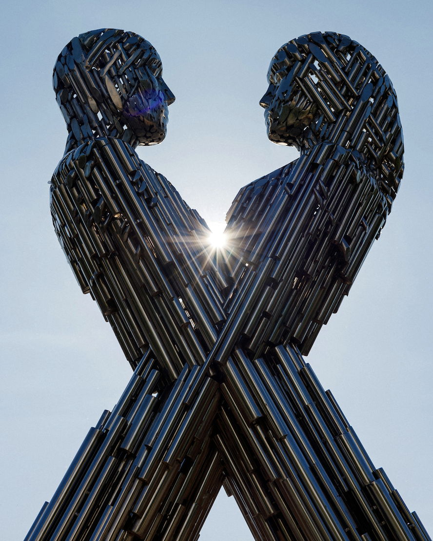 Escultura de figuras humanas hechas de tubos metálicos enfrentadas con un rayo de sol entre ellas.