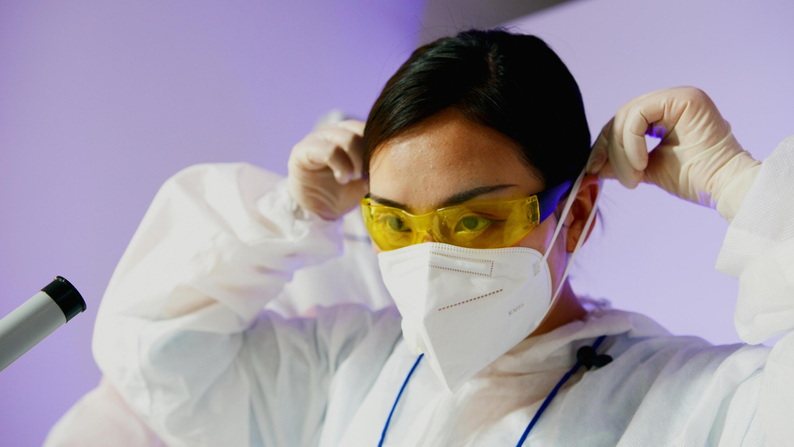 Investigadora ajusta gafas y mascarilla KN95 antes de entrar al laboratorio; protocolo de bioseguridad en biología y microbiología.