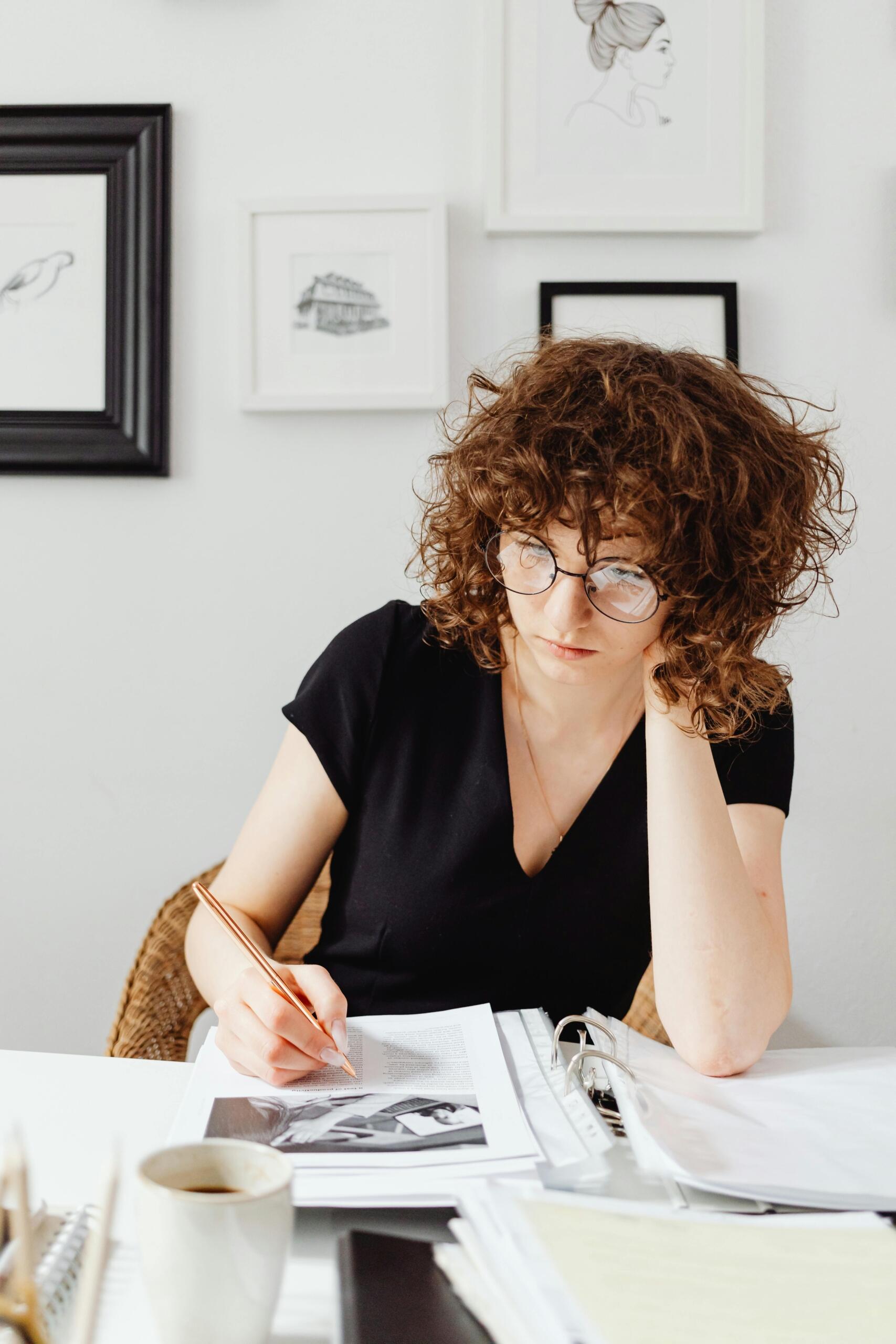 Mujer con gafas escribiendo en un cuaderno mientras estudia documentos y material de apoyo en una mesa de trabajo.