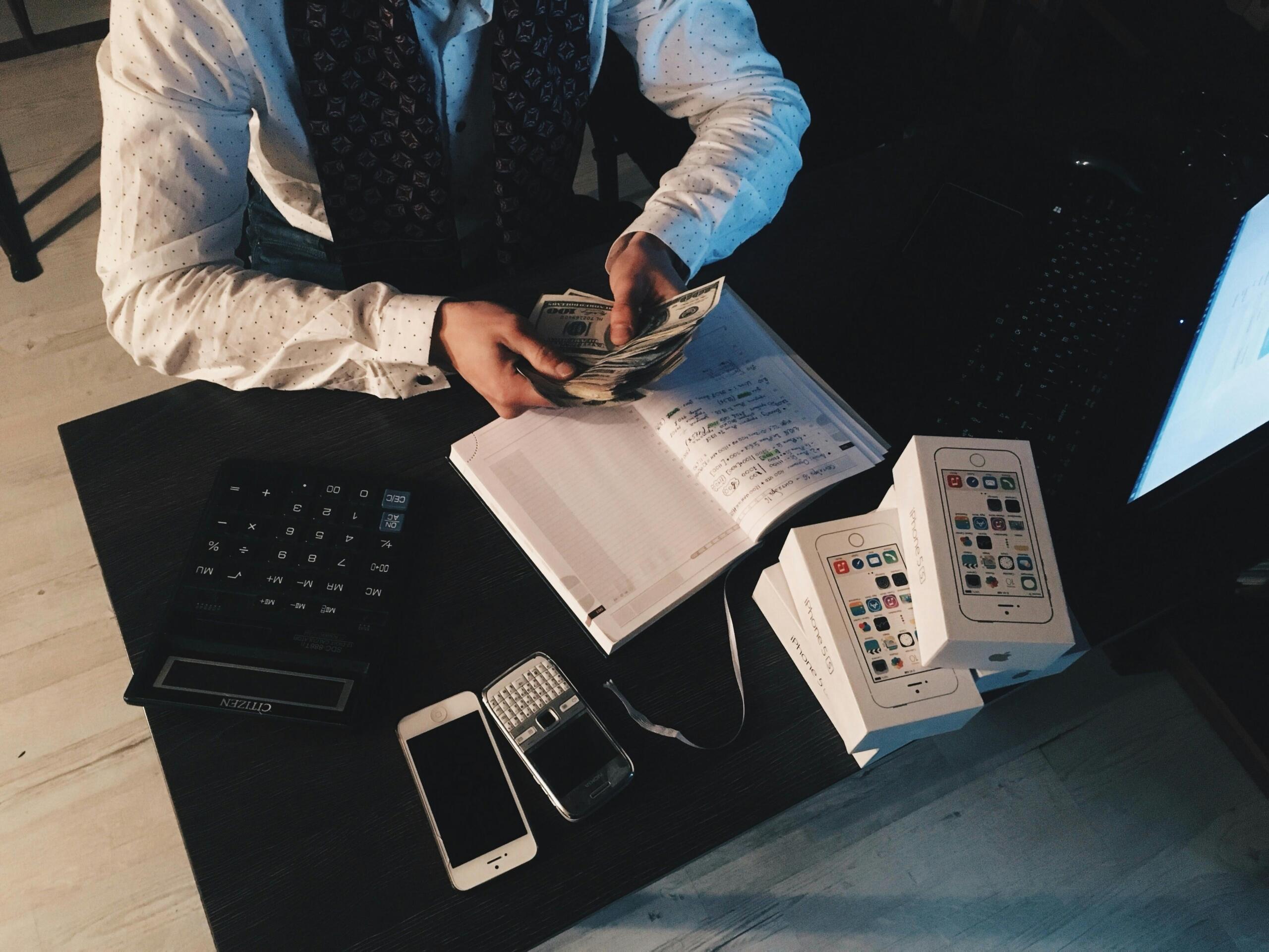 Persona sentada en un escritorio contando billetes de dólar, con calculadora, cuaderno, teléfonos y cajas de iPhone al lado.