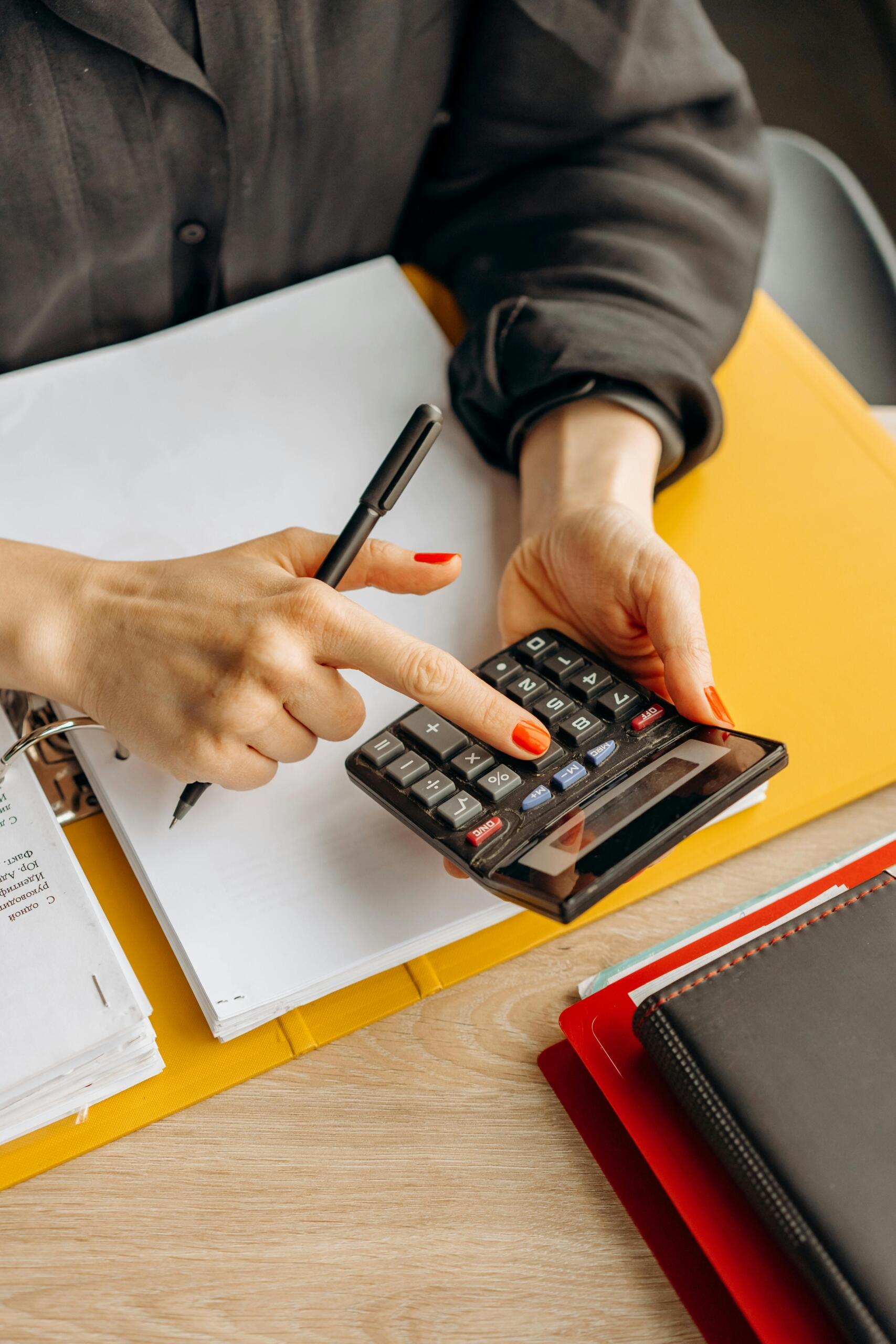 Persona usando una calculadora con una mano mientras sostiene un bolígrafo con la otra, sobre un escritorio con carpetas y documentos.
