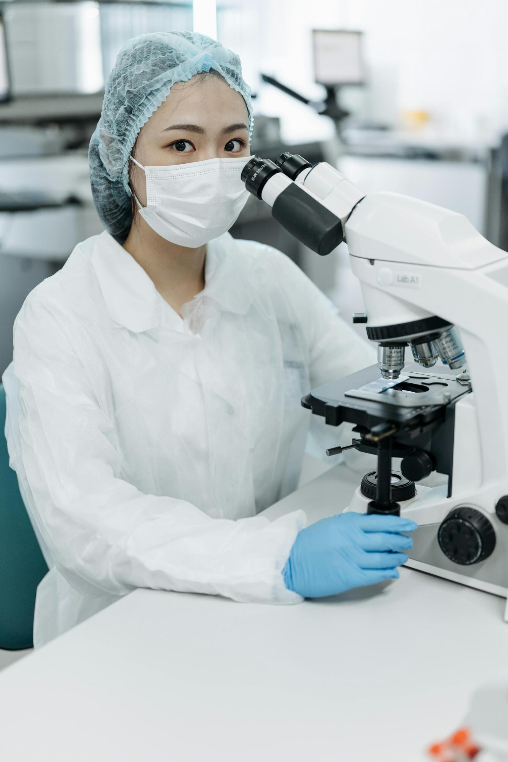 Técnica de laboratorio con bata, gorro y mascarilla opera un microscopio óptico; diagnóstico biomédico y control de calidad.