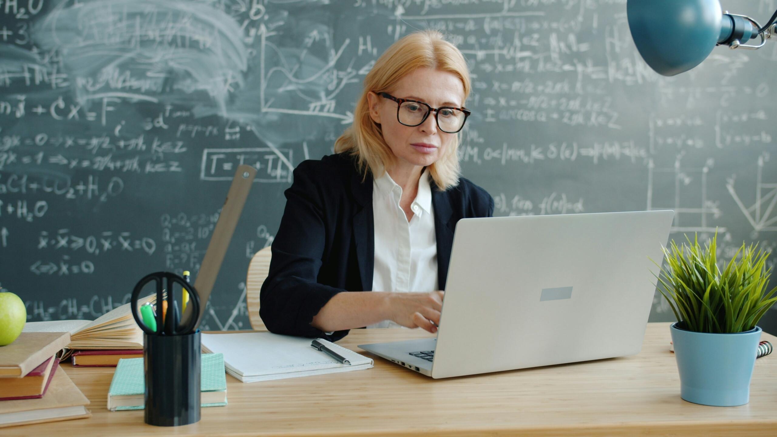 Maestra preparando material educativo en su computadora frente a una pizarra con fórmulas matemáticas, durante clases online en Panamá.
