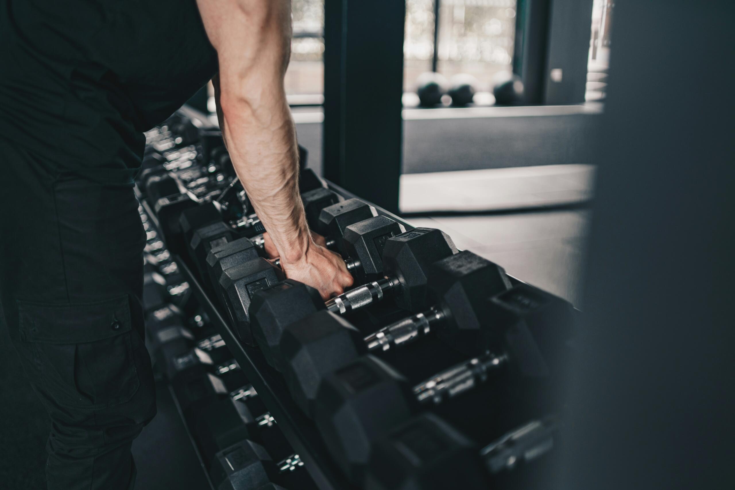 Mano seleccionando mancuernas en estante de gimnasio, preparación para entrenamiento de fuerza.