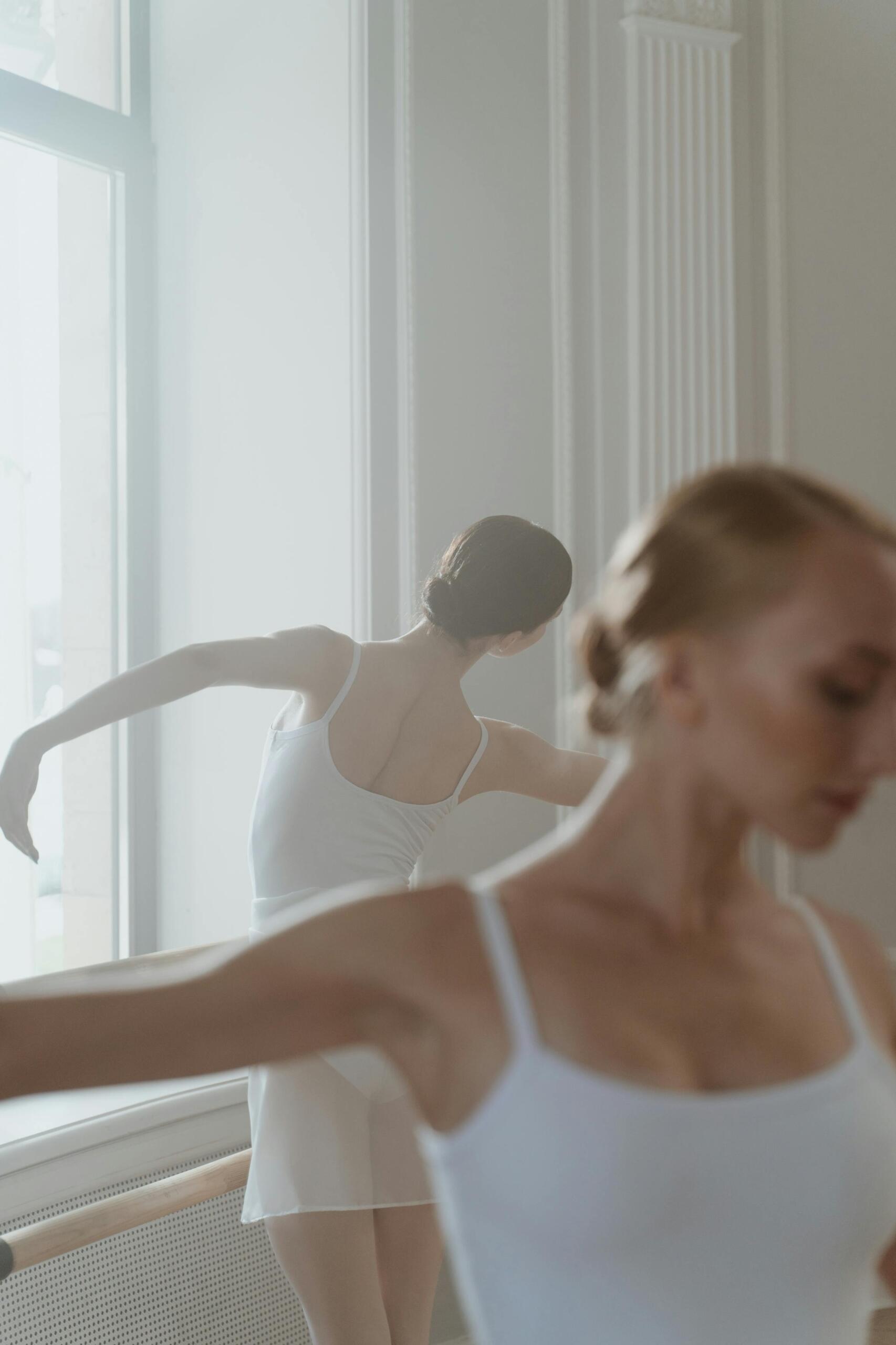 Bailarina de ballet practicando frente al espejo en un estudio luminoso, con maillot blanco y brazos extendidos.