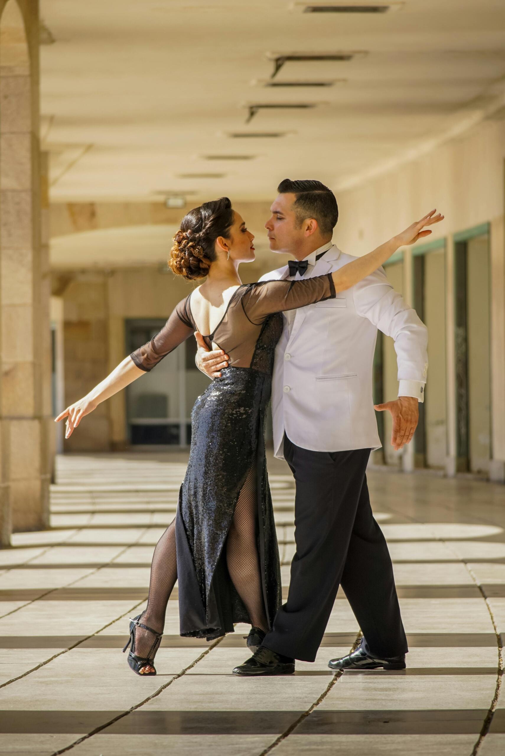 Pareja de bailarines de salón ejecutando una pose elegante en un pasillo exterior, ella con vestido negro de lentejuelas y él con esmoquin blanco.