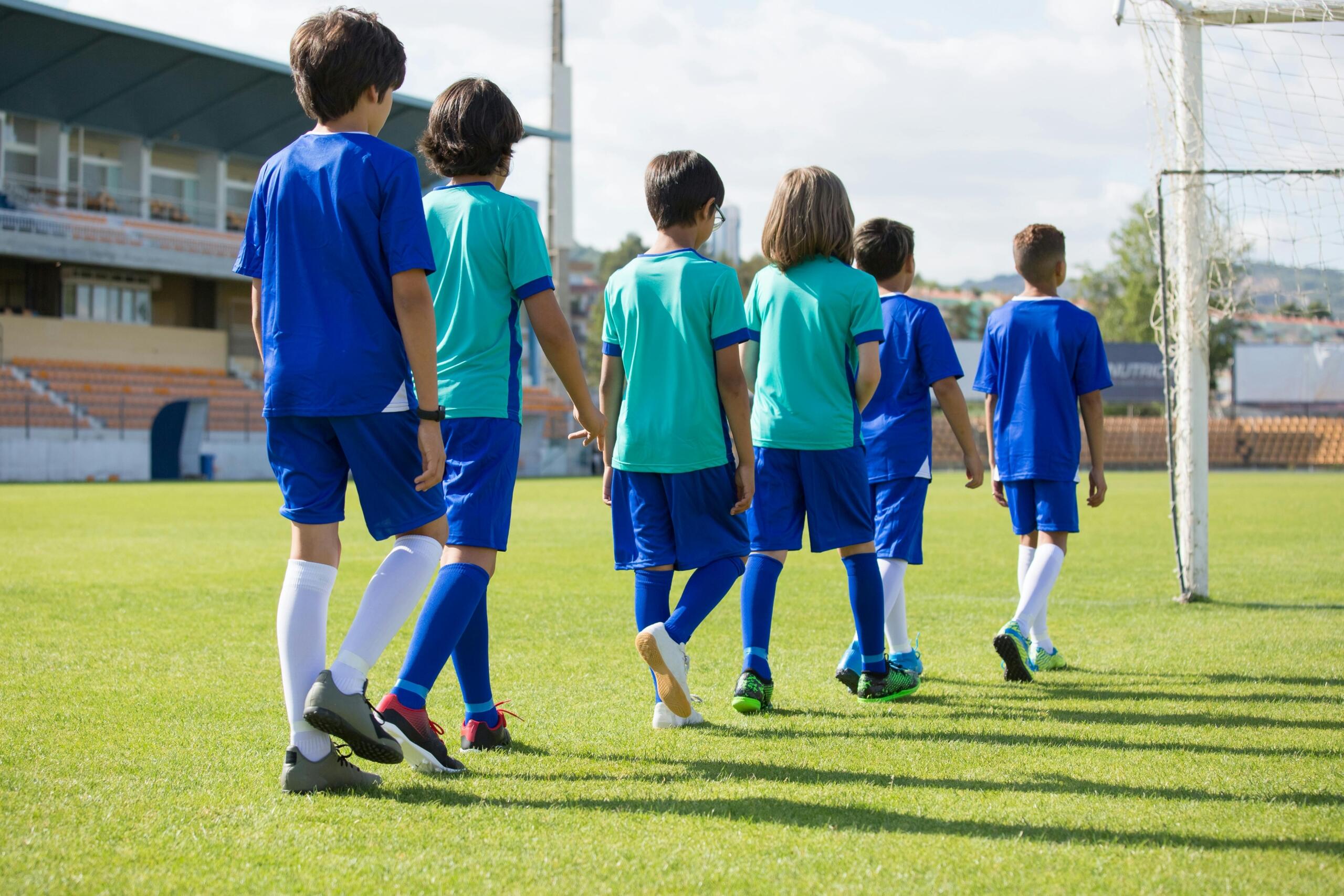 Grupo de niños con uniformes azules y turquesa caminando juntos hacia la portería en un campo de fútbol durante un entrenamiento.
