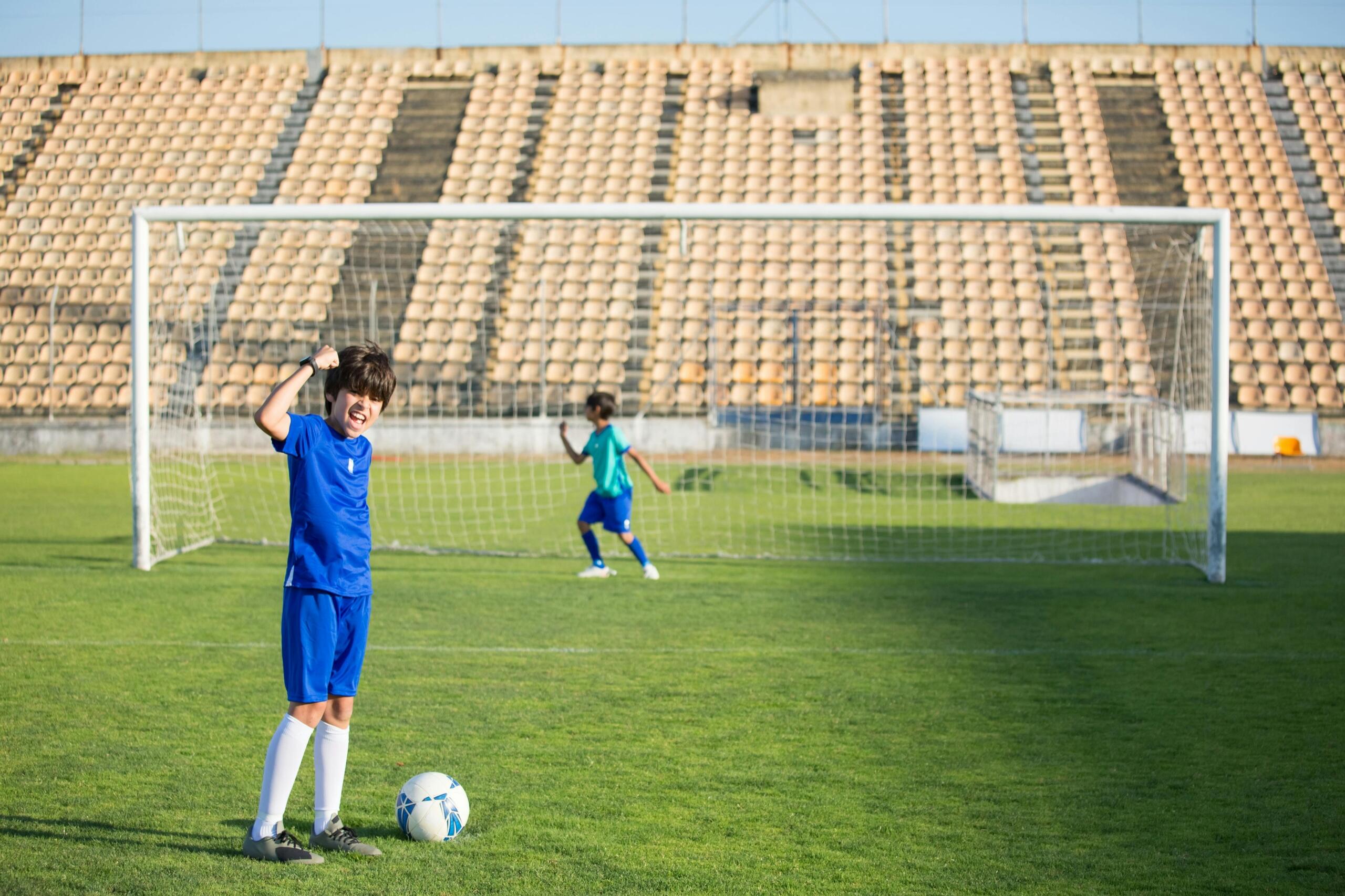 Niño con uniforme azul celebrando con entusiasmo frente a la portería mientras un compañero juega detrás de él.