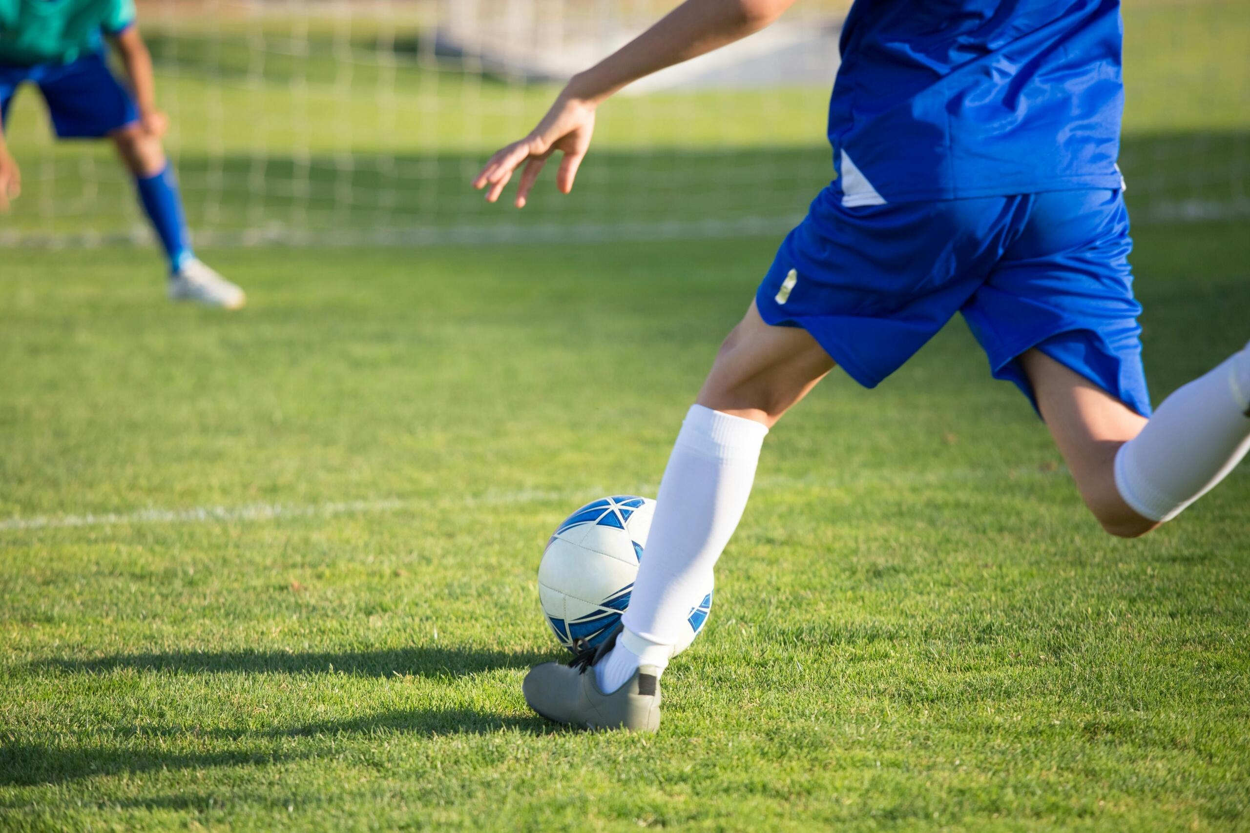 Niño con uniforme azul haciendo un tiro al balón durante una práctica de fútbol, mientras otro jugador defiende la portería.