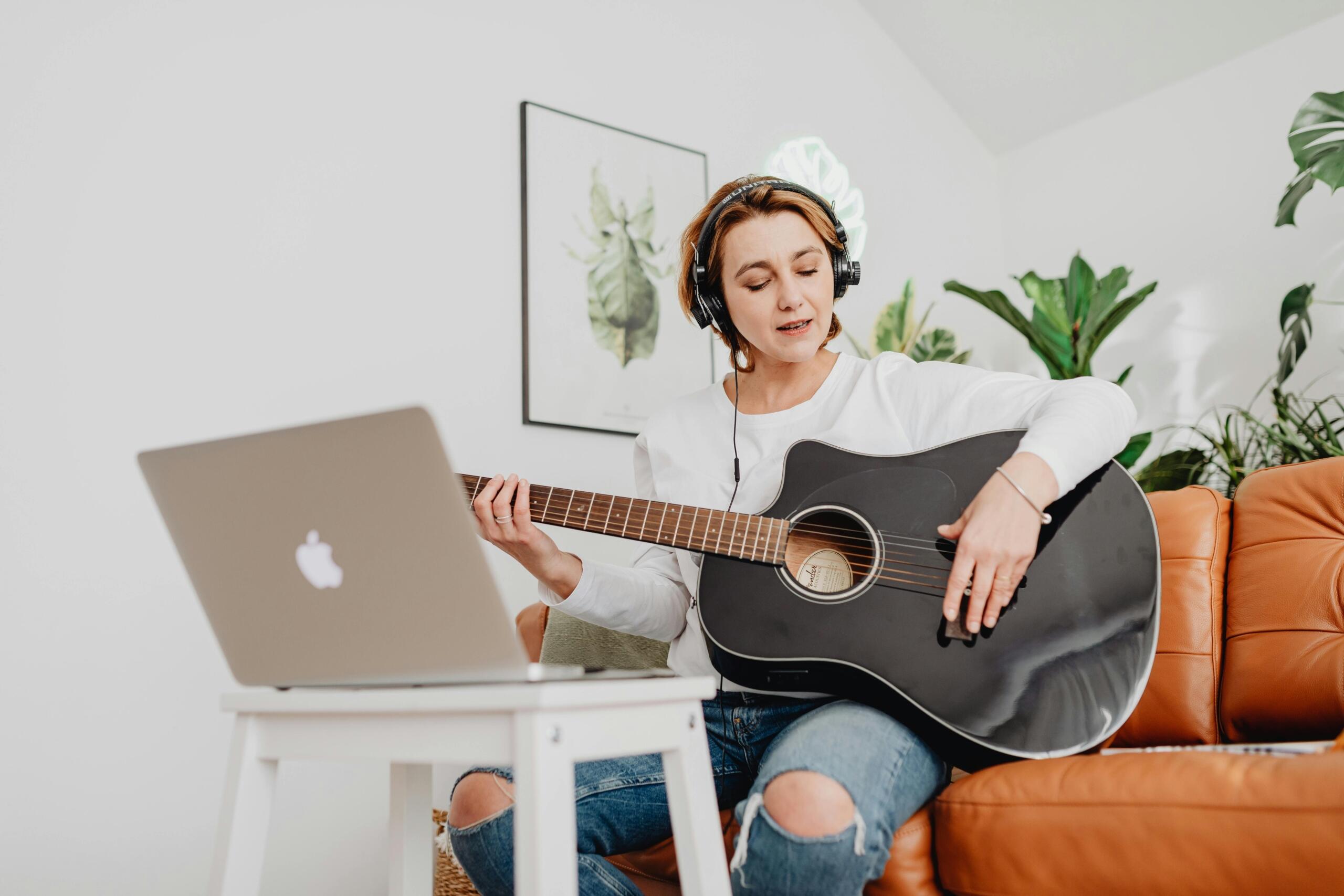 Estudiante practicando guitarra durante una clase virtual, usando audífonos y siguiendo instrucciones desde su laptop.