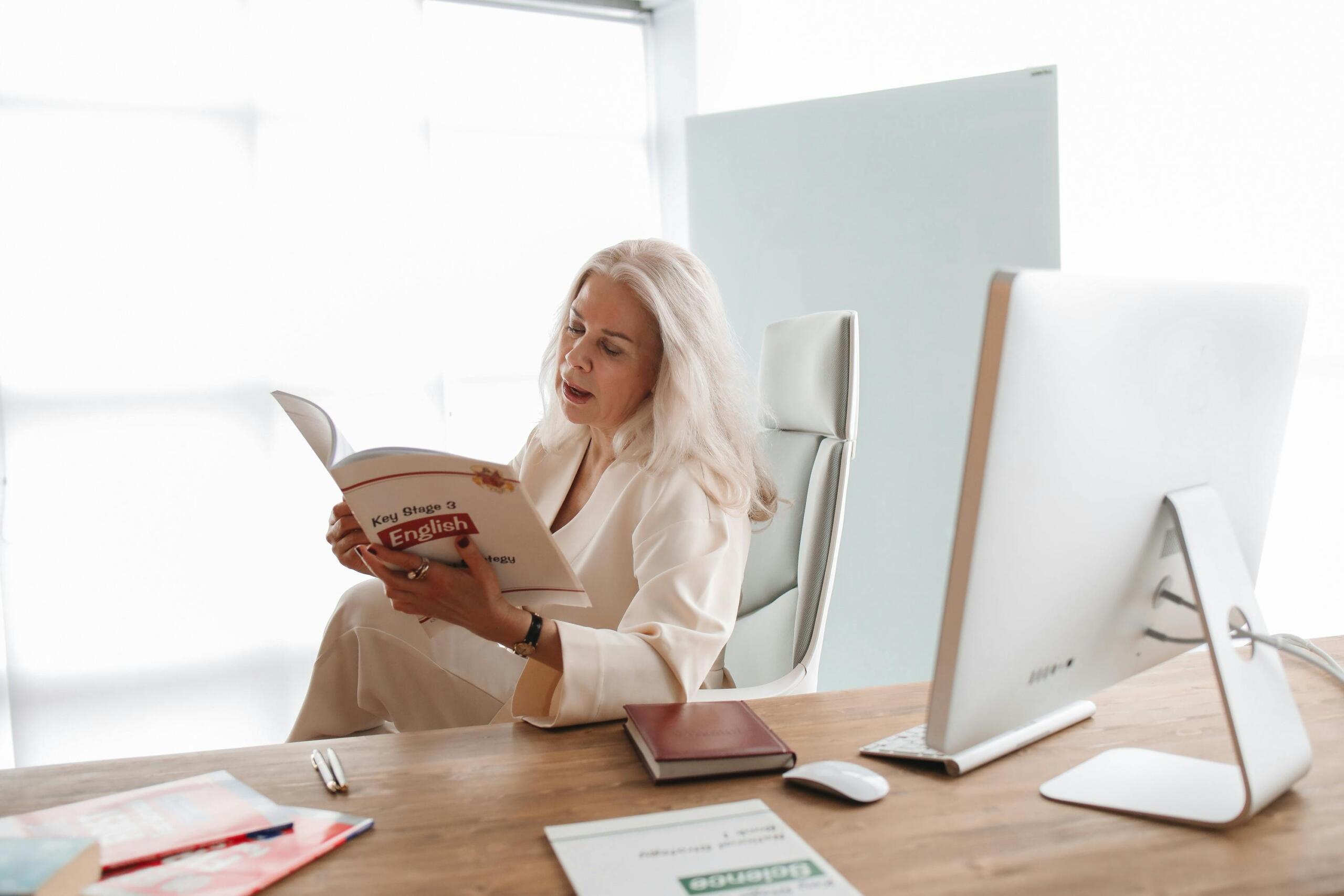 Profesora sentada en su oficina leyendo un libro de inglés frente a un monitor blanco y materiales de estudio.