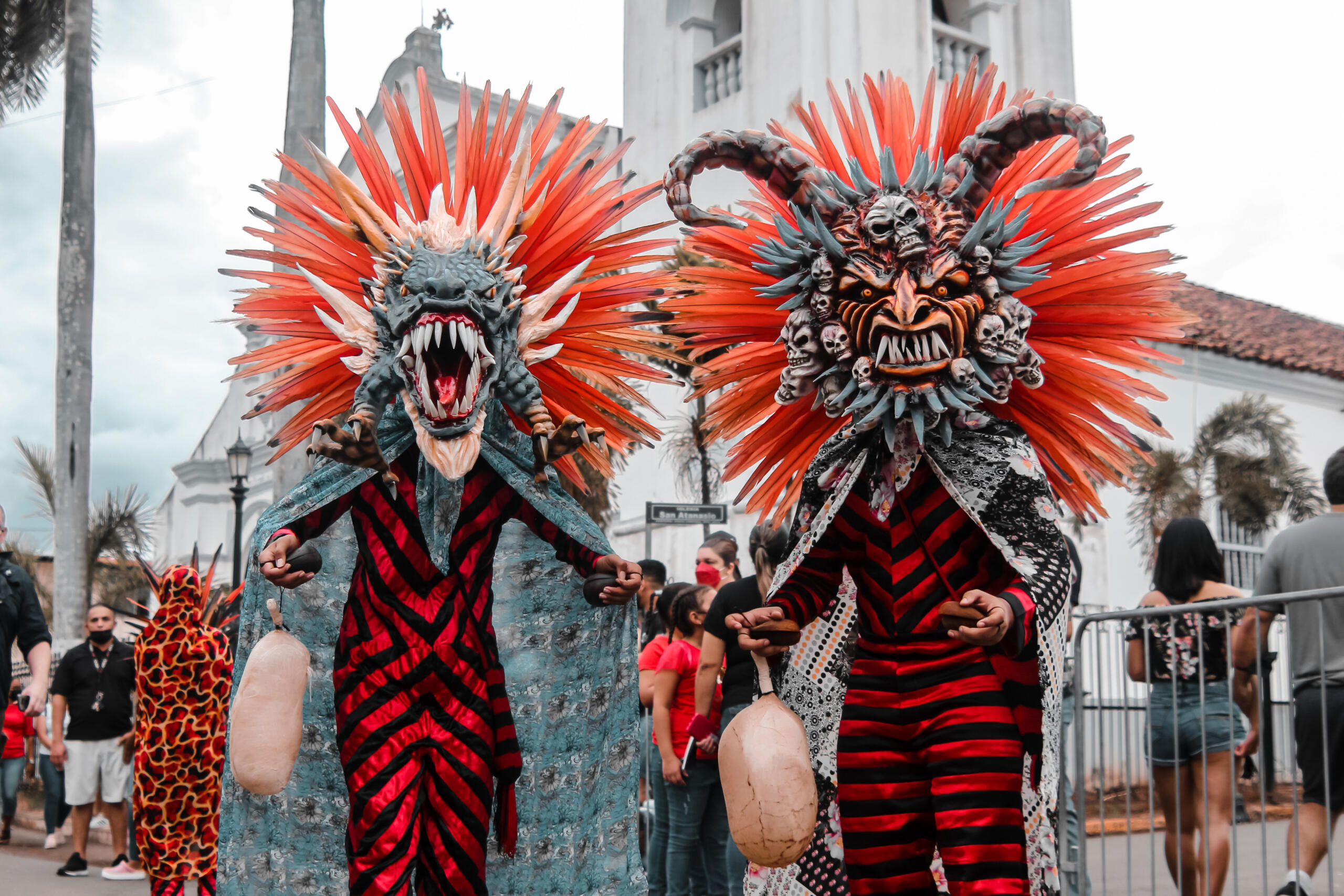 Dos diablicos sucios con máscaras elaboradas y plumas naranjas frente a iglesia colonial.
