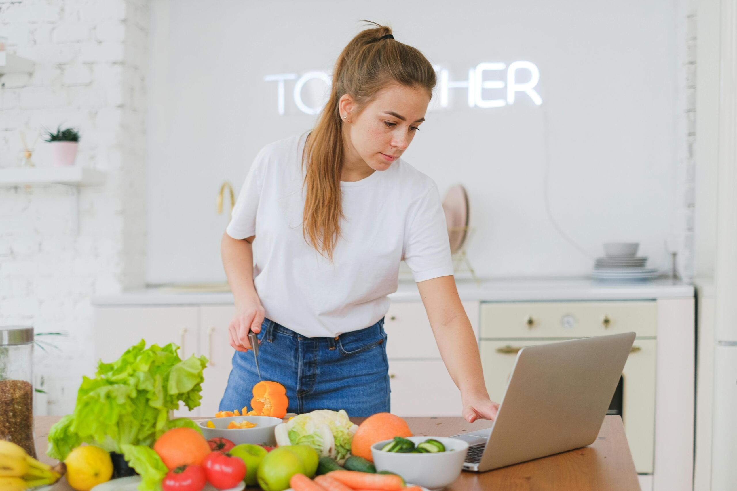Mujer aprendiendo a cocinar en casa siguiendo un canal de cocina en su laptop mientras corta verduras frescas en la cocina.