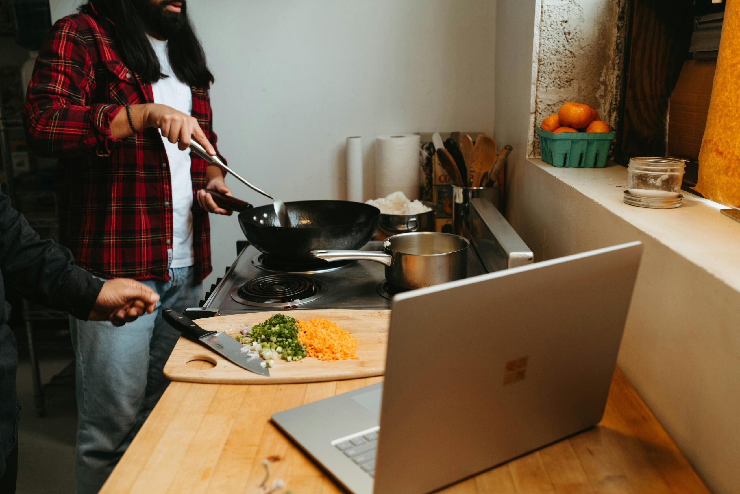 Persona cocinando en casa mientras sigue una receta en laptop como método práctico para aprender a cocinar online.
