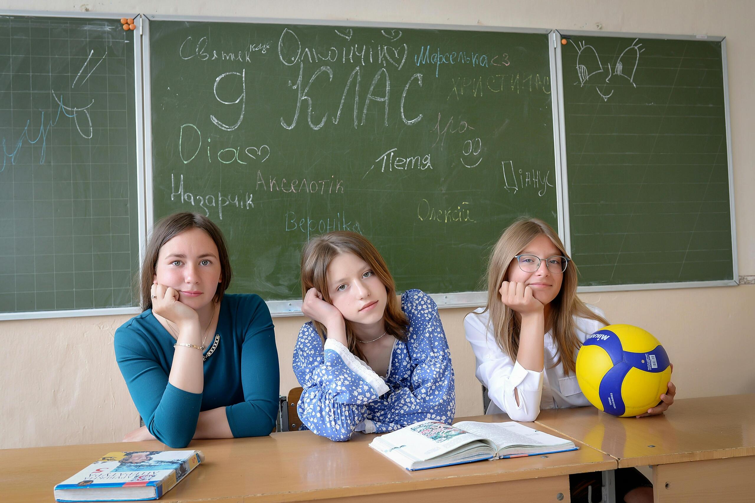 Tres estudiantes adolescentes posando aburridas en aula escolar frente a pizarra con escritura en cirílico, una sosteniendo balón de voleibol amarillo y azul.