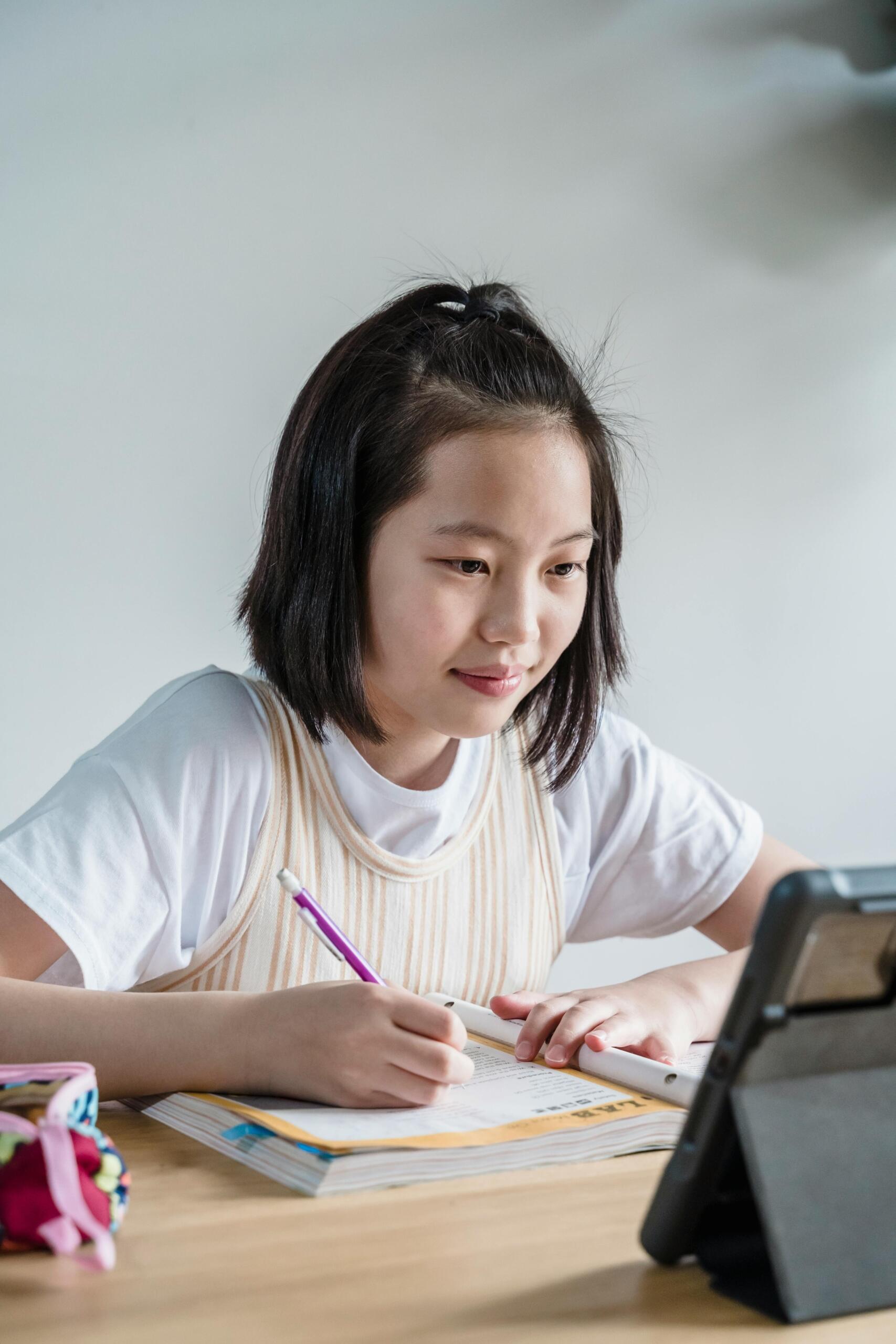 Estudiante asiática haciendo tarea con bolígrafo morado mientras consulta tableta digital, ejemplo de aprendizaje híbrido combinando libros físicos y recursos en línea.