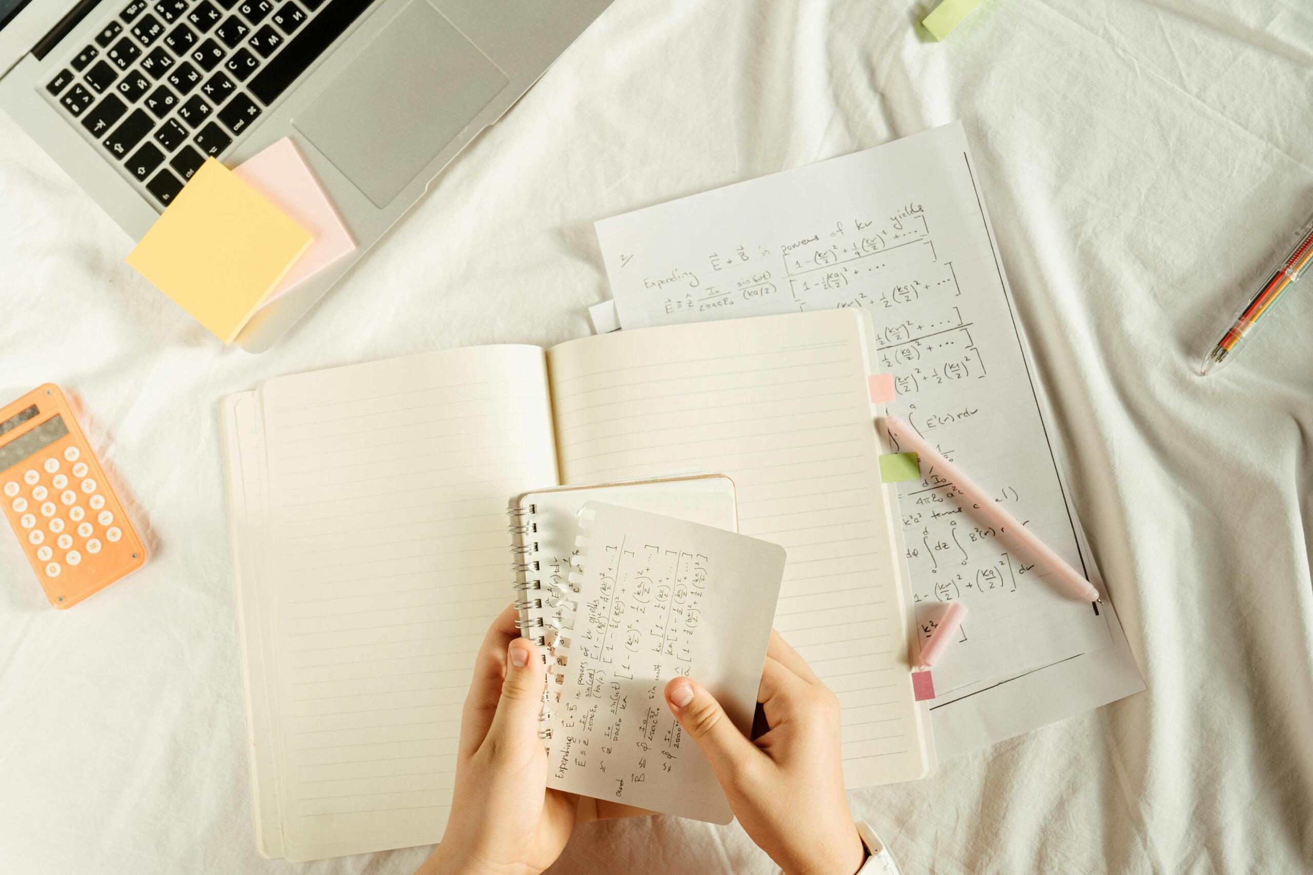Vista cenital de estudiante con cuadernos de fórmulas matemáticas, laptop, calculadora y notas adhesivas estudiando física avanzada desde casa con materiales organizados.